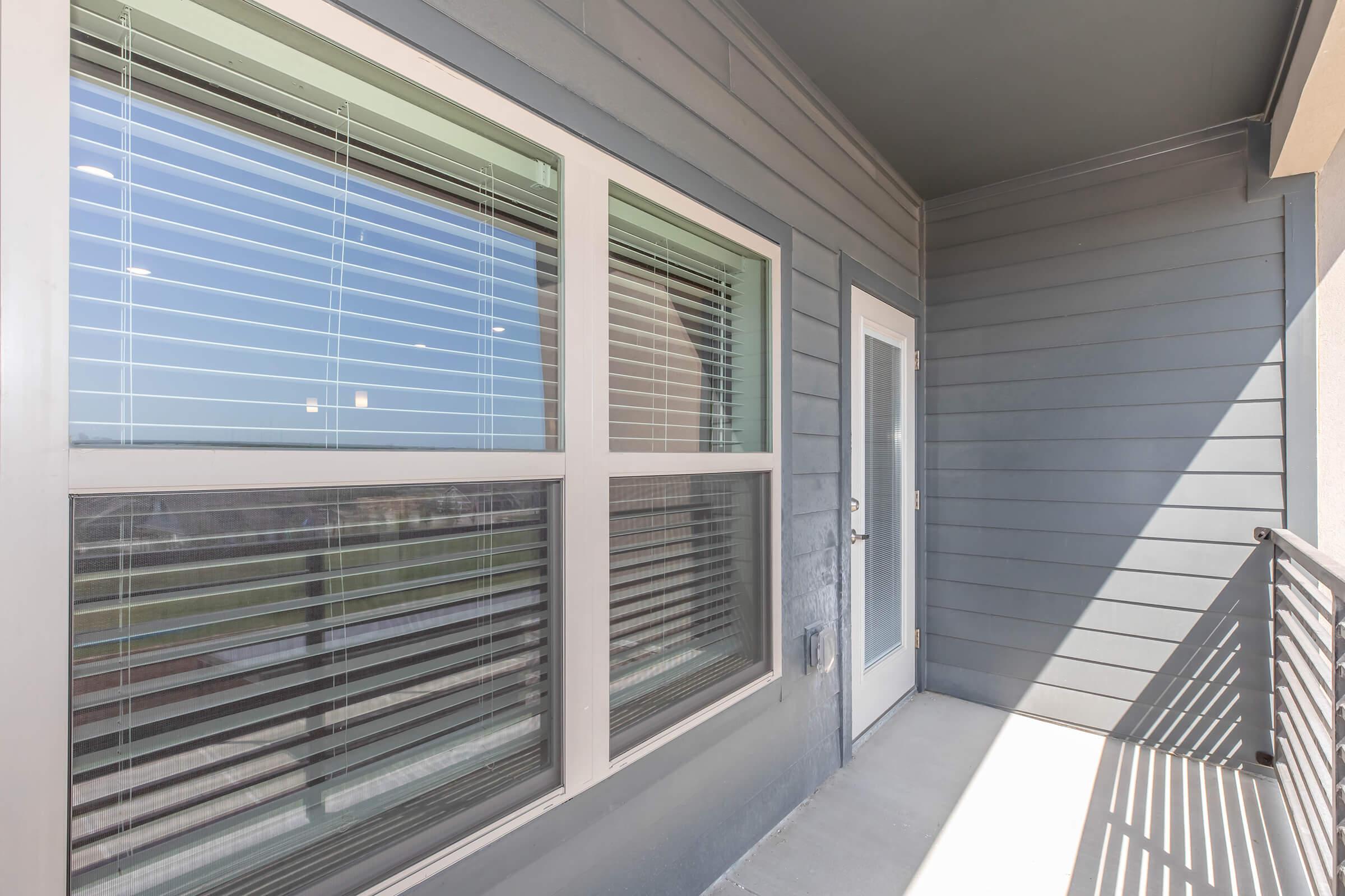 A view of a modern balcony with a gray exterior wall, featuring large windows with blinds. The door to the balcony is visible, along with a railing. Natural light is illuminating the area, and there's an open outdoor space visible beyond the balcony.