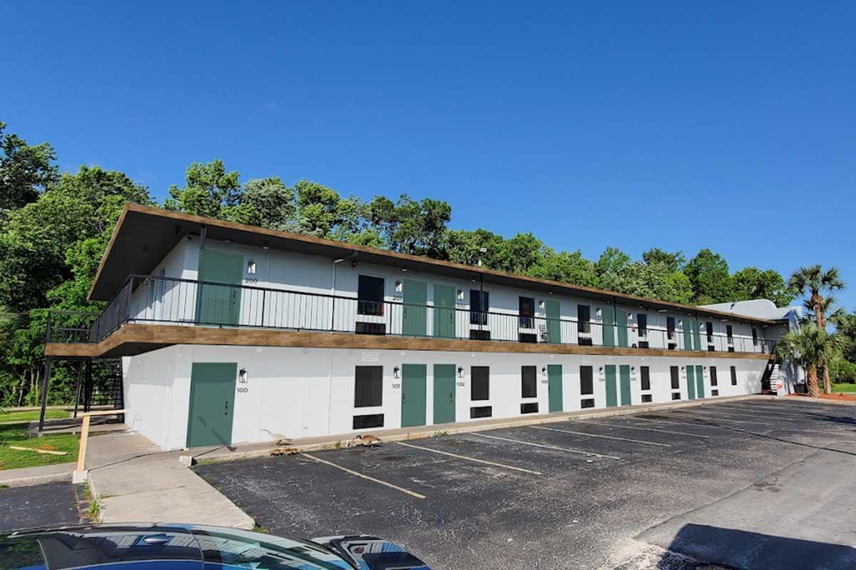 Two-story motel with green doors and balconies, surrounded by trees. The building features a parking lot in front, with several empty spaces. The sky is clear and blue, indicating a sunny day.