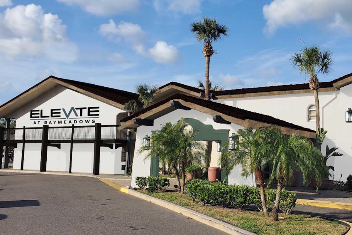 Exterior view of Elevate at Baymeadows, a building featuring a modern design with a large sign. Lush greenery and palm trees surround the entrance, while a clear blue sky with clouds is visible above. The setting has a welcoming and spacious appearance, ideal for a community or residential complex.