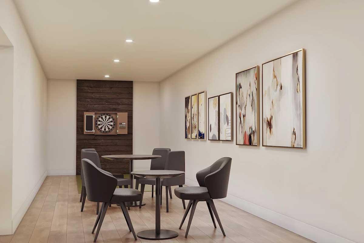 A modern lounge area featuring a small round table with four stylish chairs. On the walls, there are several framed abstract artworks. A dartboard is mounted on a wooden accent wall, creating a cozy and inviting space for relaxation and socializing. The flooring is sleek and wooden.