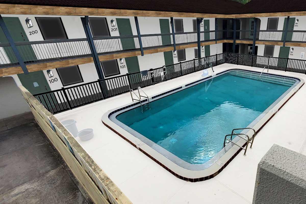 A view of a courtyard with a swimming pool surrounded by two-story motel buildings. The pool features a shallow and deep end, with ladders for access. Each building has numbered doors, and the area is enclosed with railings. Sunlight reflects on the water surface, creating a bright atmosphere.