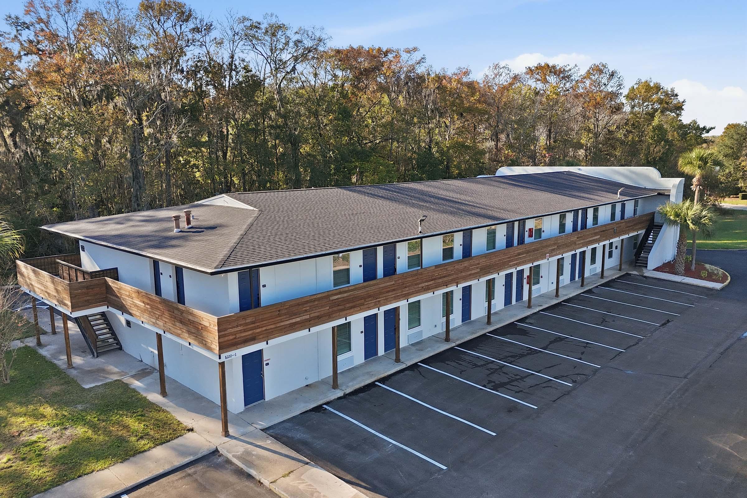 A two-story motel building with blue doors and wooden accents, surrounded by greenery. Several parking spaces are visible in front, with a clear blue sky above and trees in the background showcasing autumn foliage. The scene conveys a peaceful, inviting atmosphere.