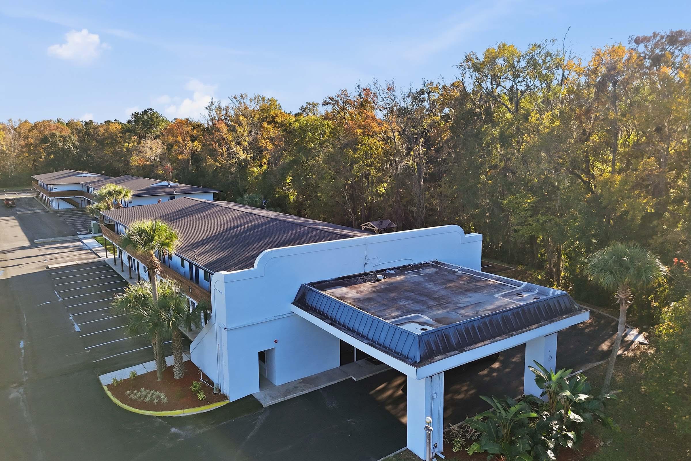 Aerial view of a blue building with a flat roof, surrounded by a paved parking lot and lush trees with autumn foliage. The building features a covered entrance and well-maintained landscaping, set against a clear, blue sky.