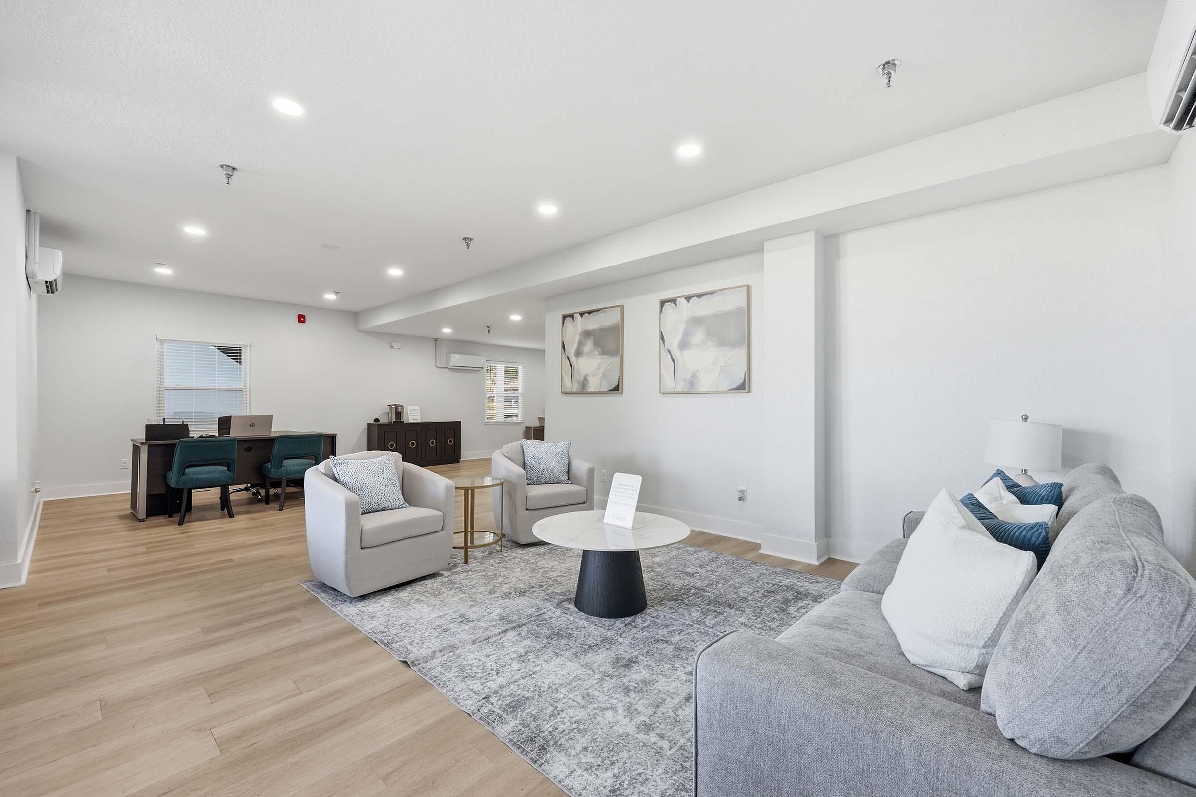 A modern living room featuring two light grey armchairs and a grey sofa, accented with decorative pillows. A round black coffee table sits on a light-colored rug. The space has wood flooring and a desk area with two chairs in the background, complemented by abstract wall art and natural light from a window.