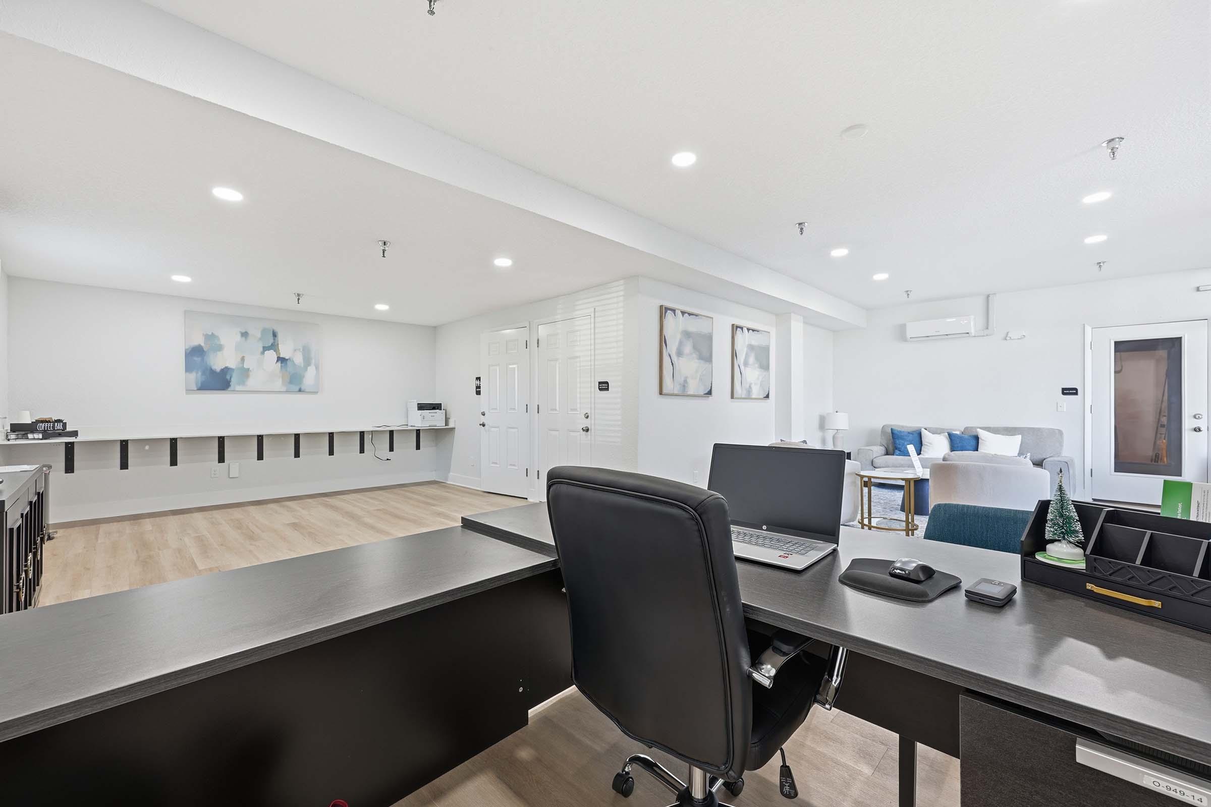 A modern office space featuring a black desk with a laptop and chair, facing a light-colored couch and decorative wall art. The room has bright lighting, white walls, and an open layout with additional workspace visible in the background.