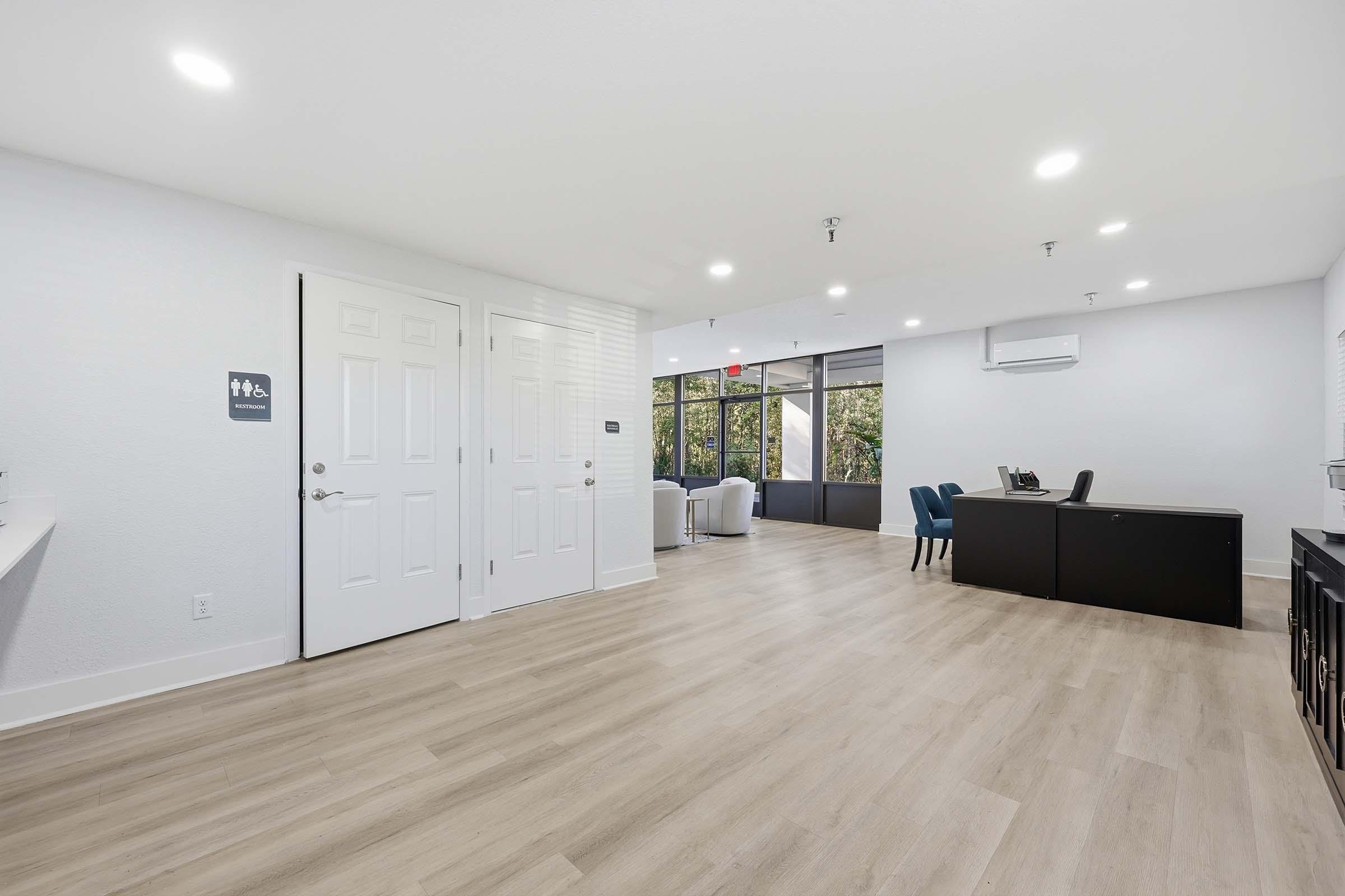 A spacious, modern office or waiting area with light wood flooring, large windows allowing natural light, and contemporary furniture. It includes a desk and chairs, a sign indicating restrooms, and a minimalist design with white walls and ceiling lights.