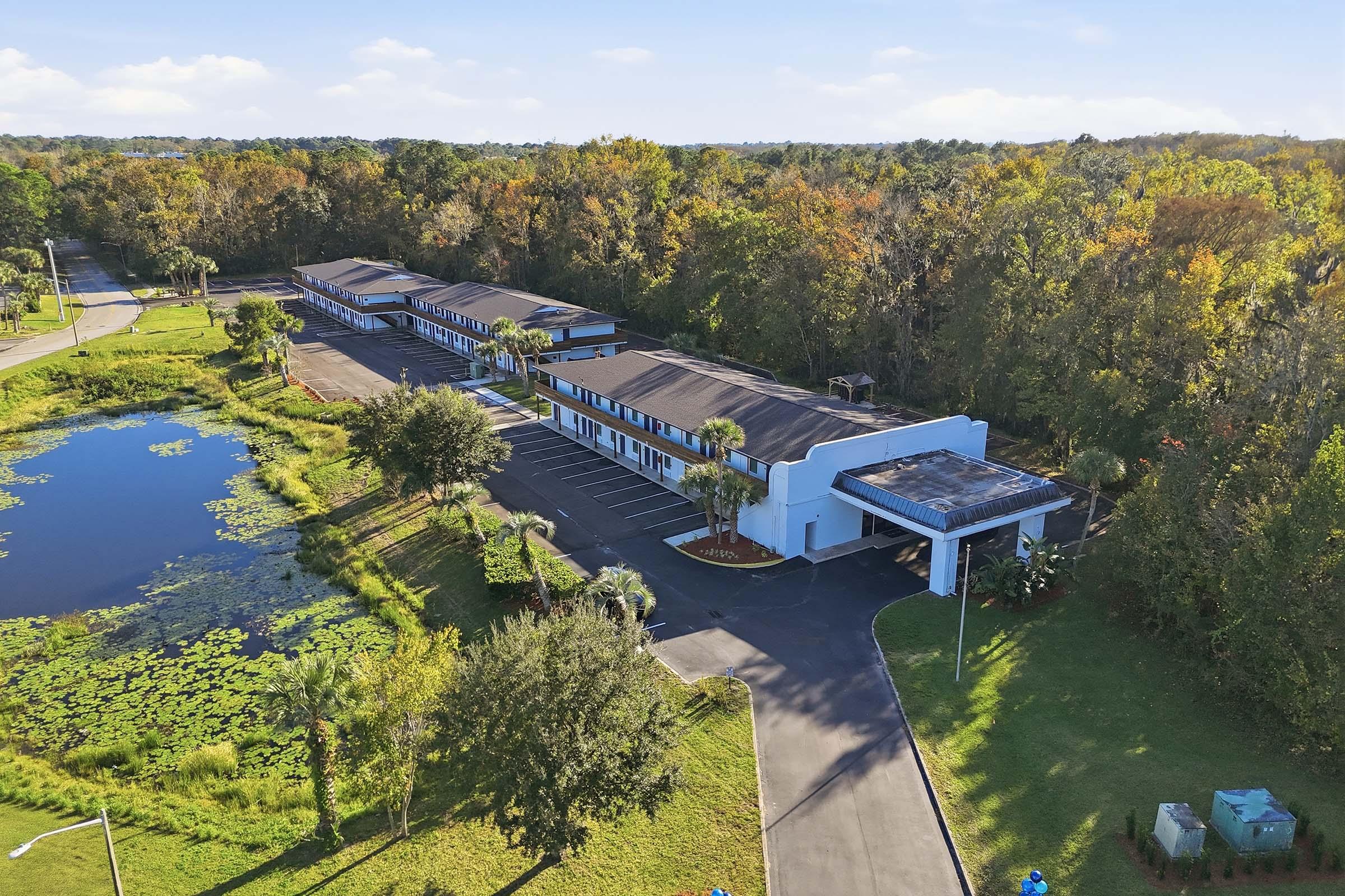 Aerial view of a low-rise motel situated near a pond, surrounded by greenery. The building features multiple rooms with a parking lot in front, landscaped with palm trees. The scene showcases clear skies and autumn foliage in the background.
