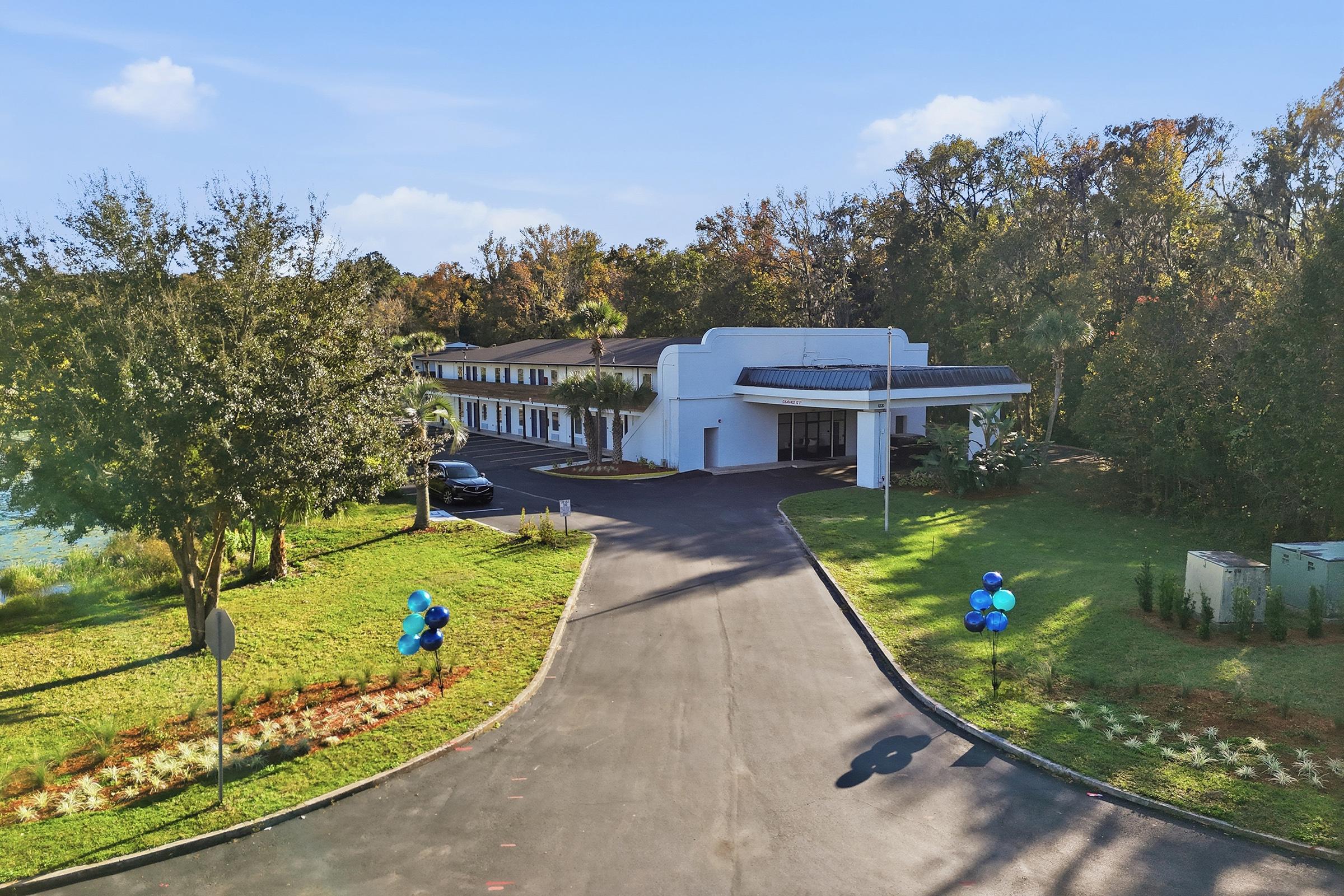 A modern building surrounded by trees and greenery, with a clear blue sky above. A paved driveway leads to the entrance, where decorative blue balloons are placed on either side. The scene suggests a welcoming atmosphere, possibly for an event or a new opening.