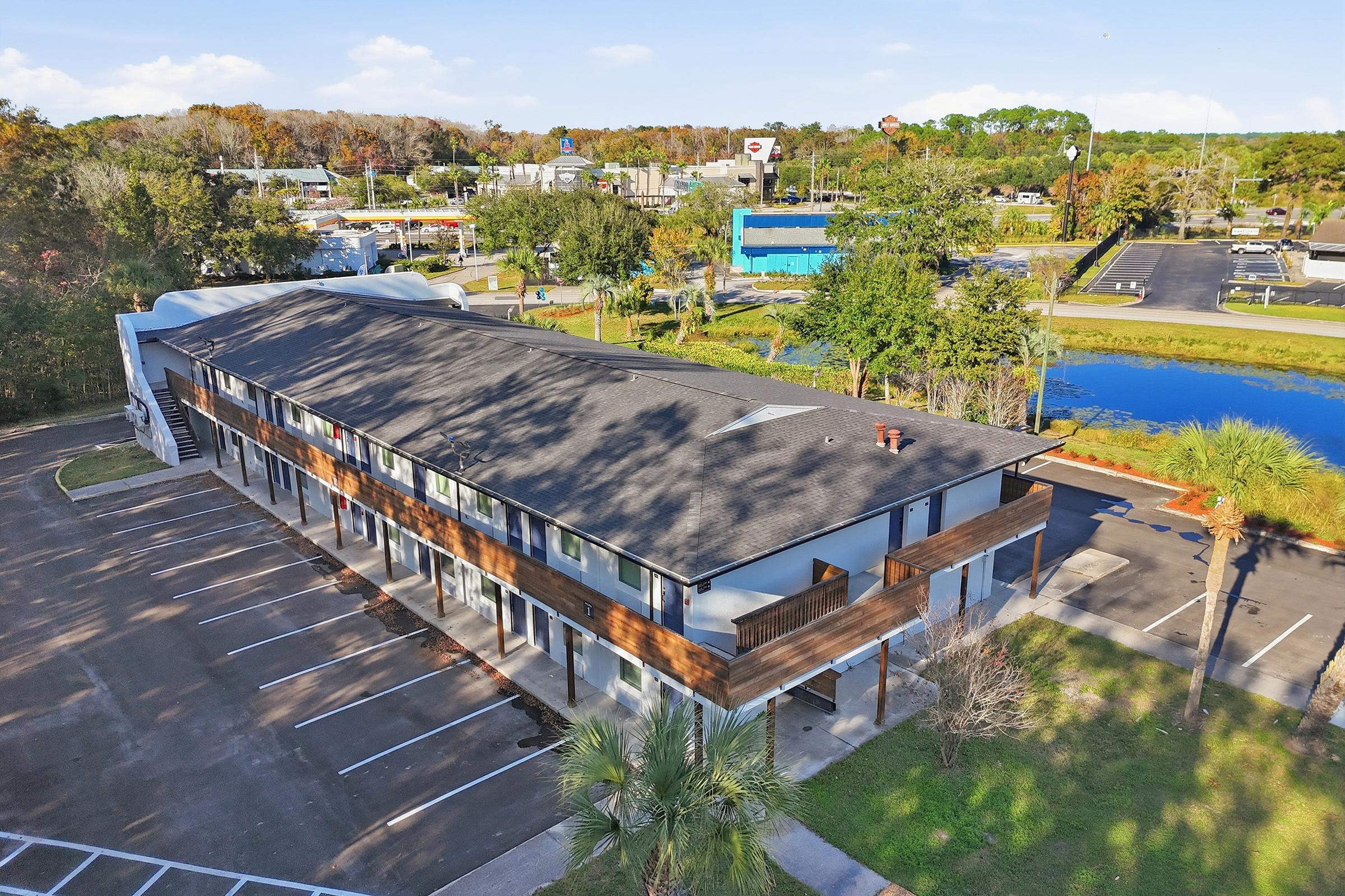 Aerial view of a modern, two-story building with a flat roof, featuring wooden accents. The surrounding area includes a parking lot with multiple spaces and palm trees, along with a small pond and greenery in the background. Several commercial buildings are visible further back.