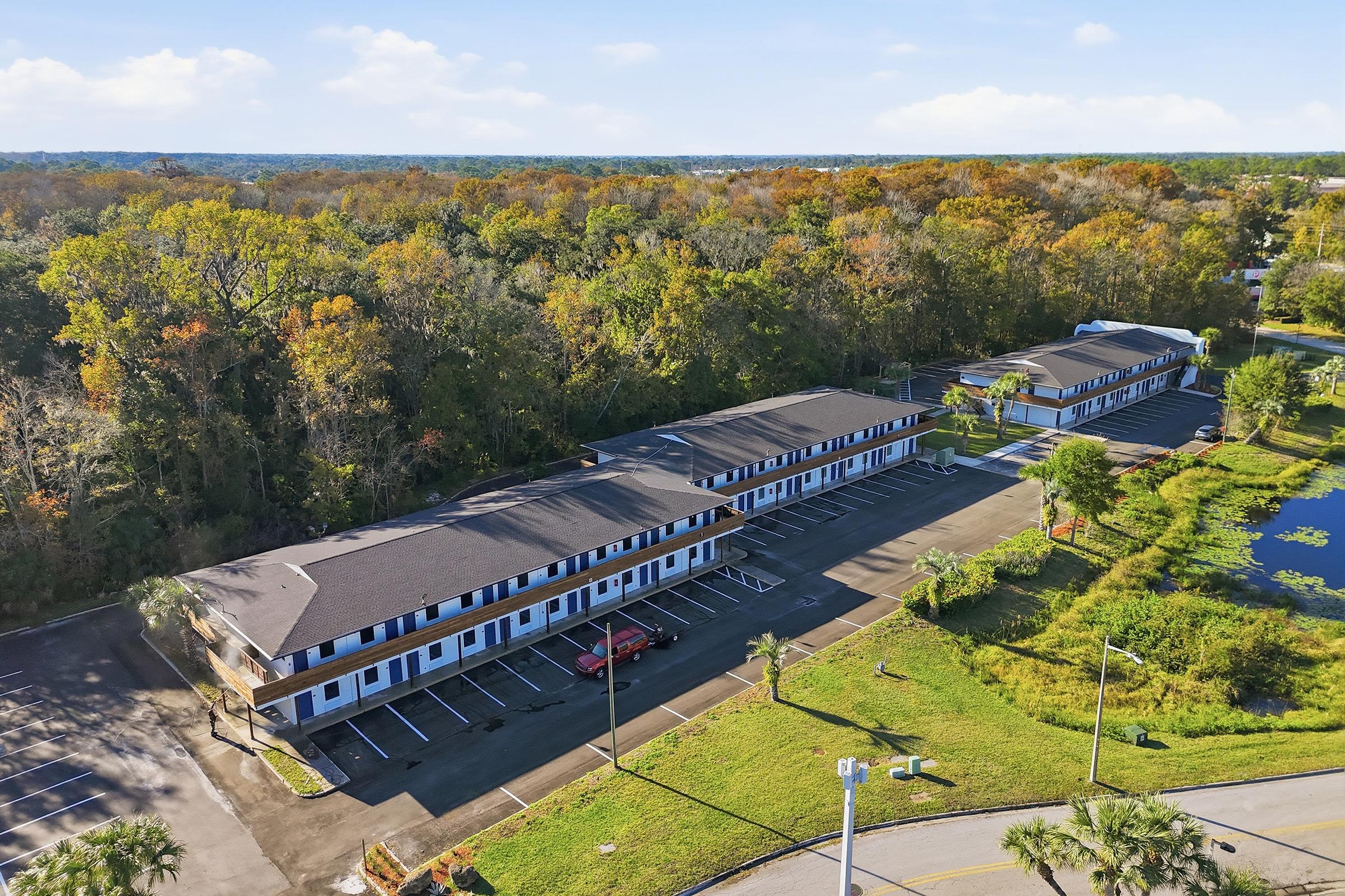 Aerial view of a multi-story motel surrounded by trees and greenery. The parking lot is mostly empty, with a few cars visible. In the background, rolling hills can be seen. The scene captures a peaceful, suburban setting with a clear blue sky.