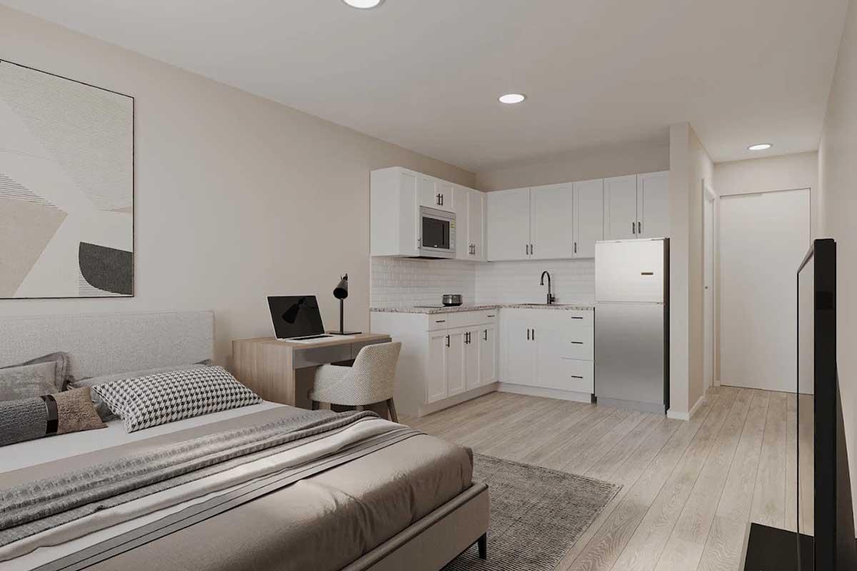 A modern studio apartment featuring a bed with neutral bedding, a small desk and chair, and a kitchen area with white cabinets, a microwave, and a stainless steel refrigerator. The room has light-colored wooden flooring and minimalistic decor, creating a clean and spacious atmosphere.