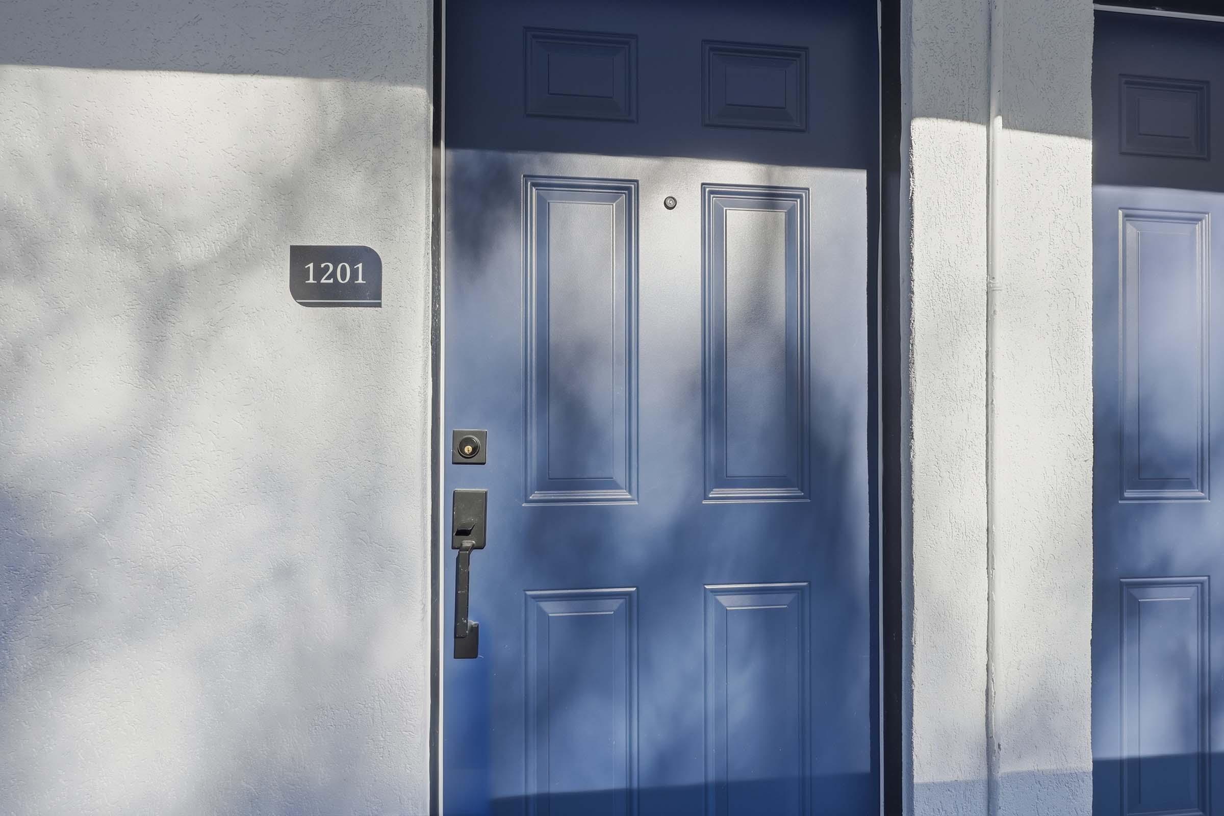 A blue door with the number 1201 displayed beside it, set against a light-colored wall. The door features a modern handle and is partially illuminated, casting soft shadows from nearby light.