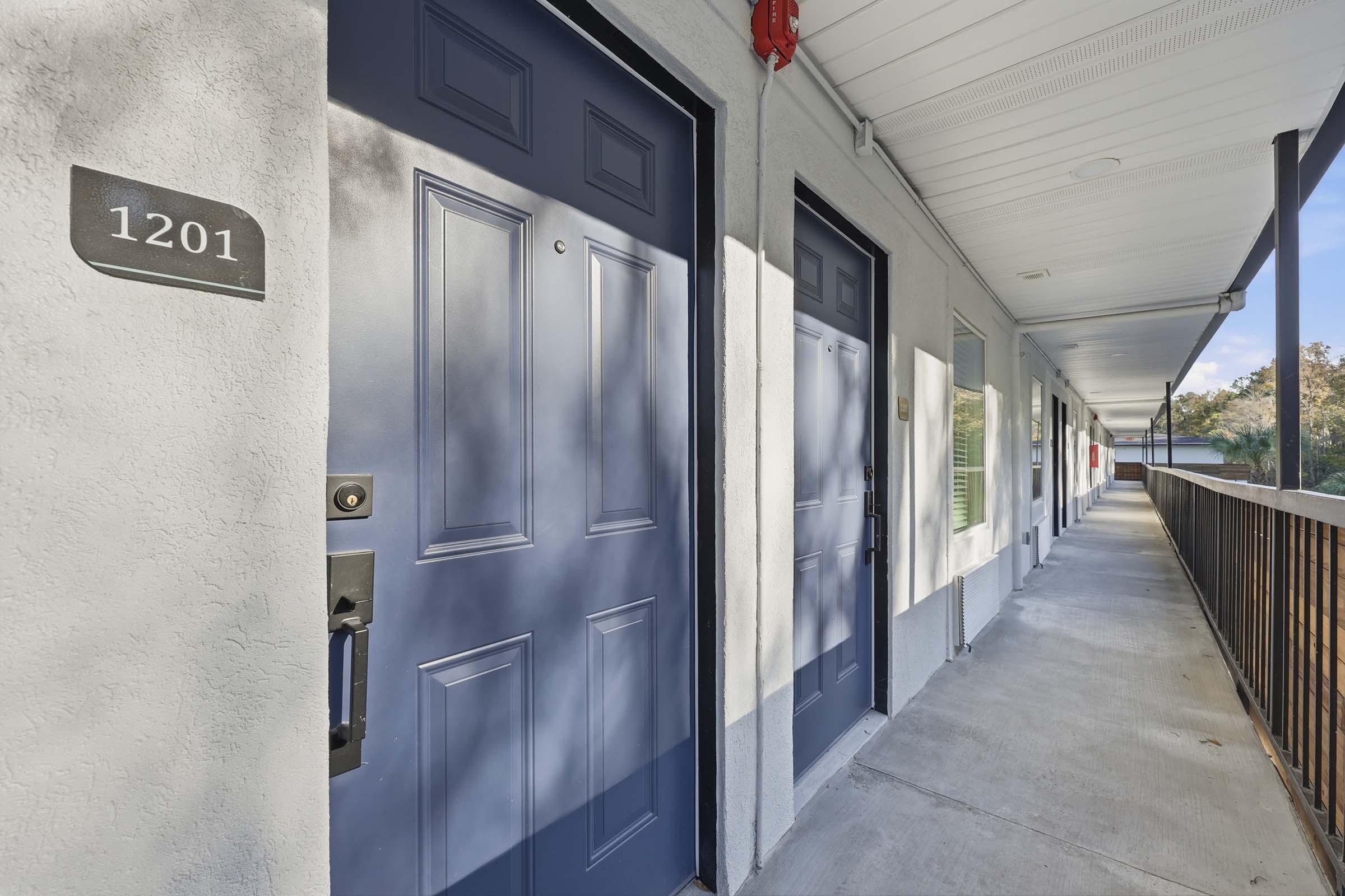 Two hotel room doors on a corridor with grey walls and a concrete walkway. The door on the left is labeled "1201" and features a modern handle. Sunlight casts shadows on the ground, and the area is surrounded by a railing.