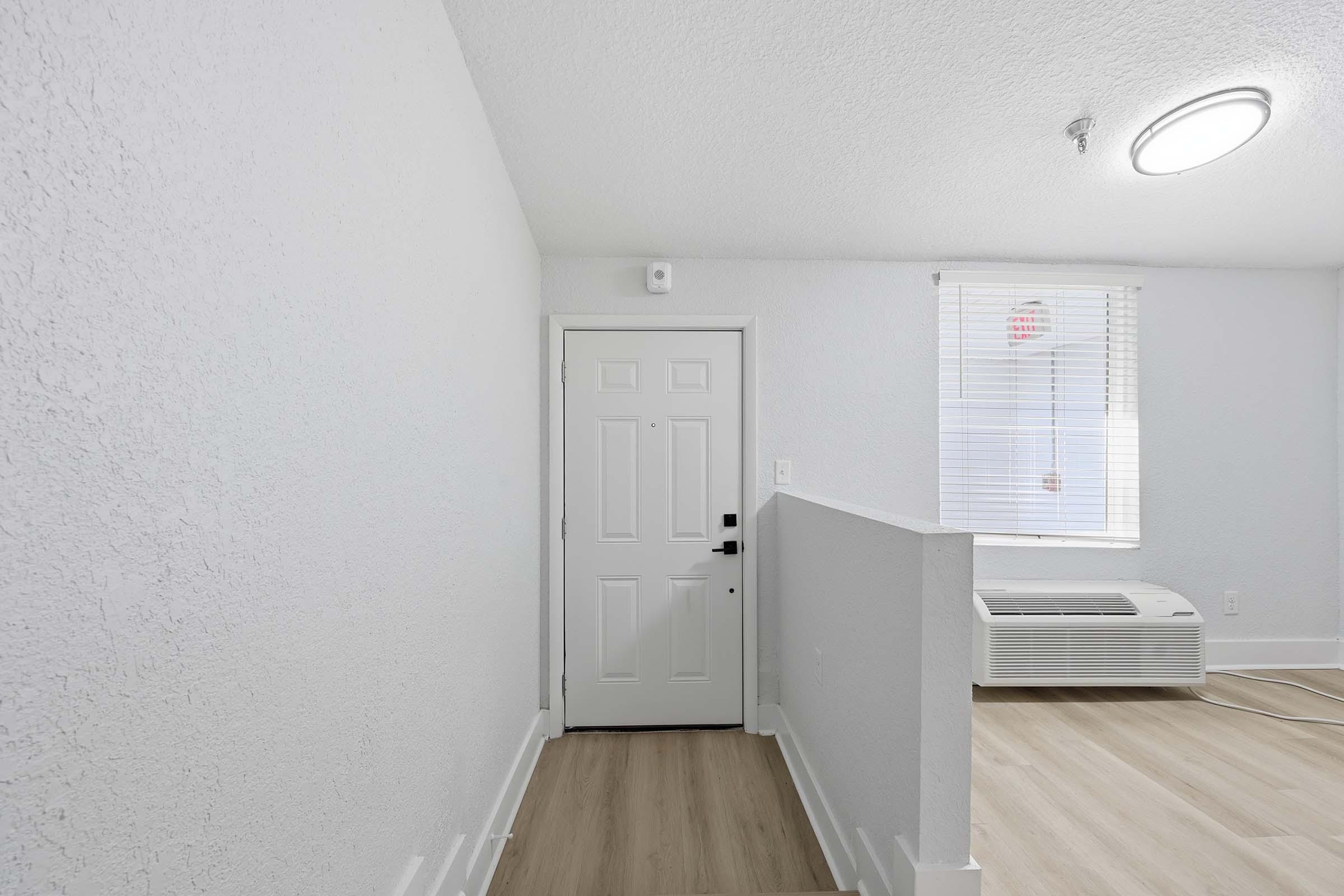 A well-lit entryway featuring a white wall, a closed door with a simple design, and a window with blinds. The floor is covered in light wood laminate, and there is a wall-mounted air conditioning unit visible in the corner. The overall decor is minimalist and modern.