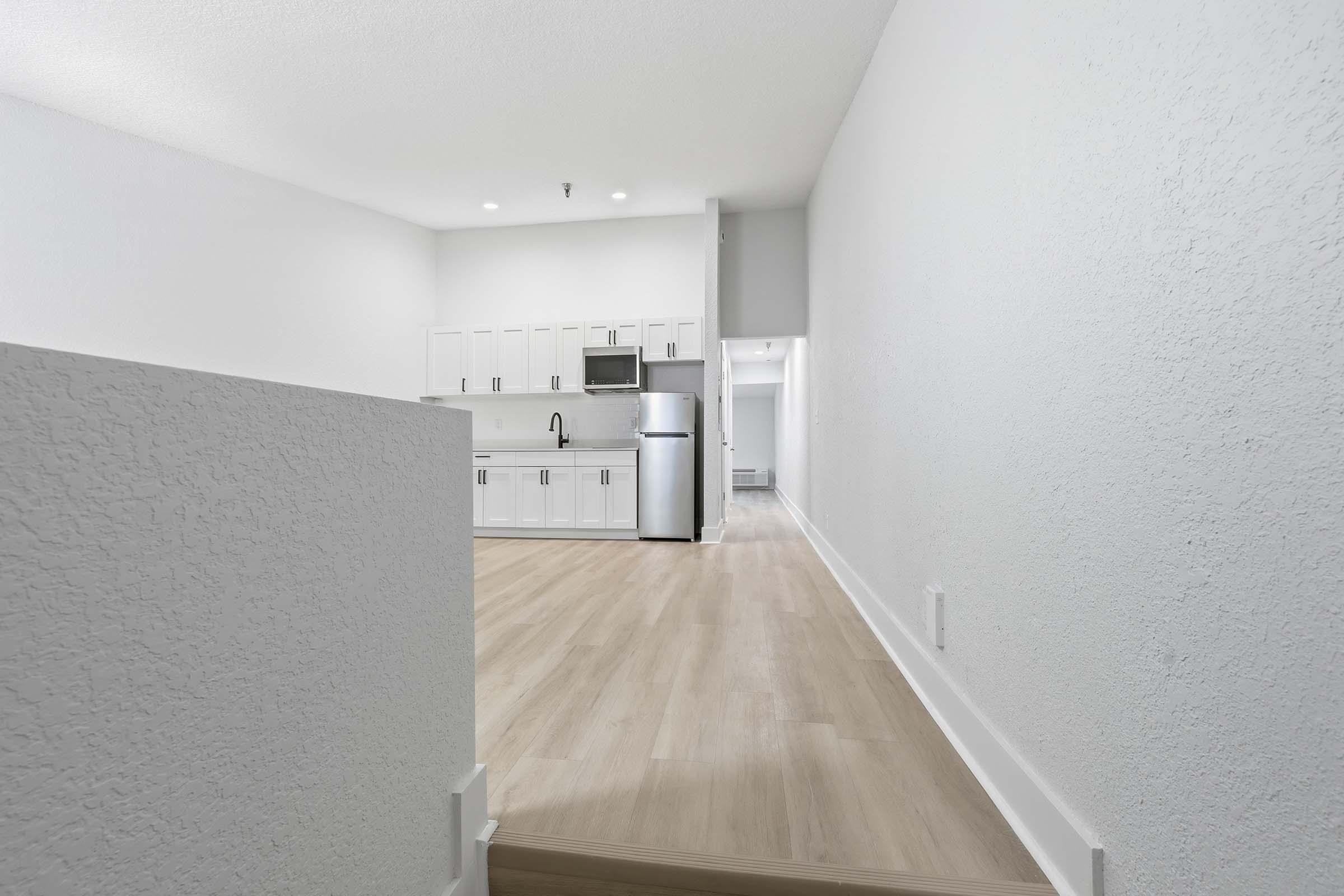 A bright, modern interior view of an apartment showing a light wood floor leading to a kitchen with white cabinets, a stainless steel refrigerator, and a sleek countertop. The walls are painted in a light color, creating a spacious feel, and there is a hallway visible in the background.