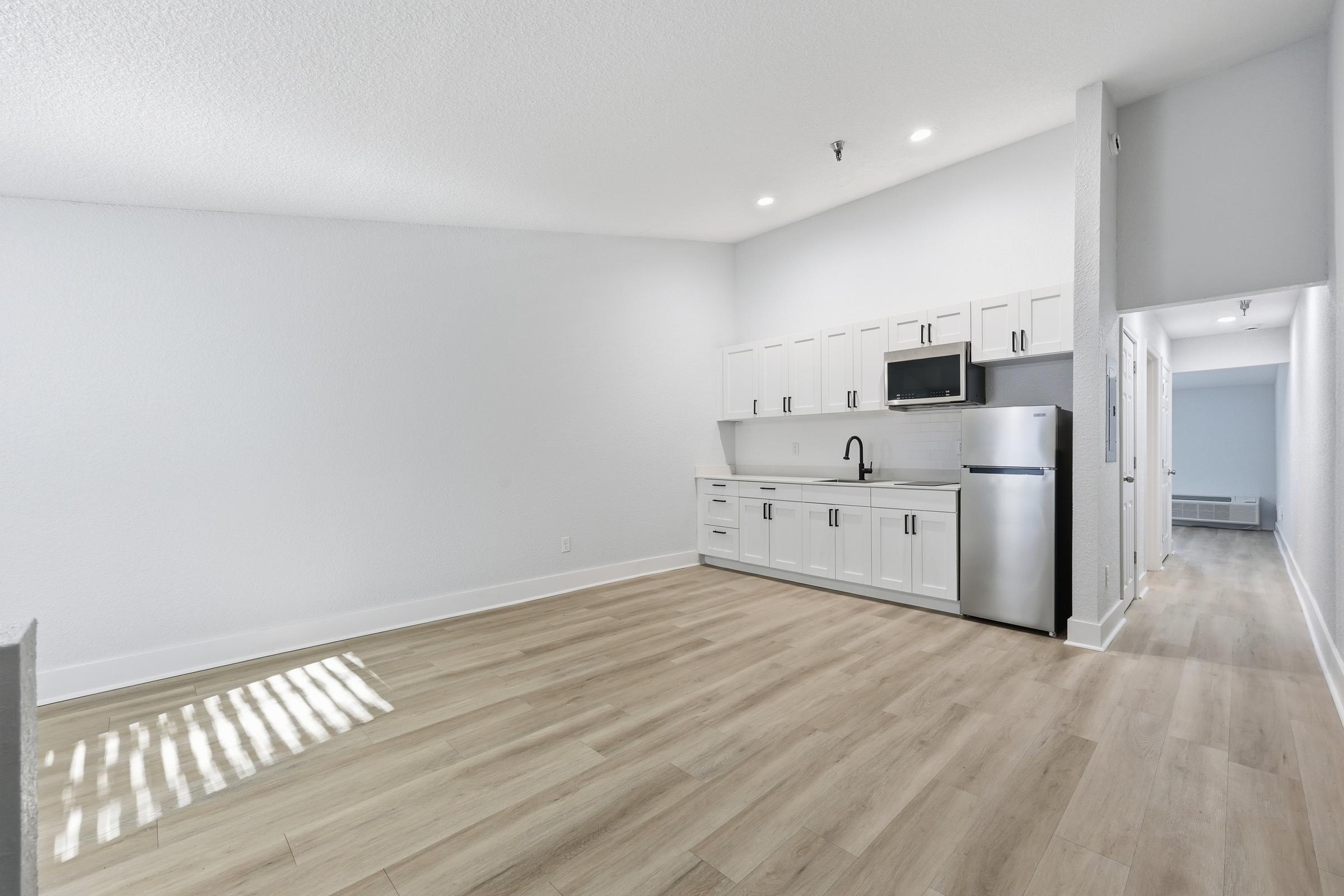Bright and modern studio apartment with white walls and light wood flooring. Features a compact kitchen area with white cabinets, a microwave, and a stainless steel refrigerator. Sunlight streams in through a window, enhancing the spacious feel. A hallway leads to other rooms in the background.