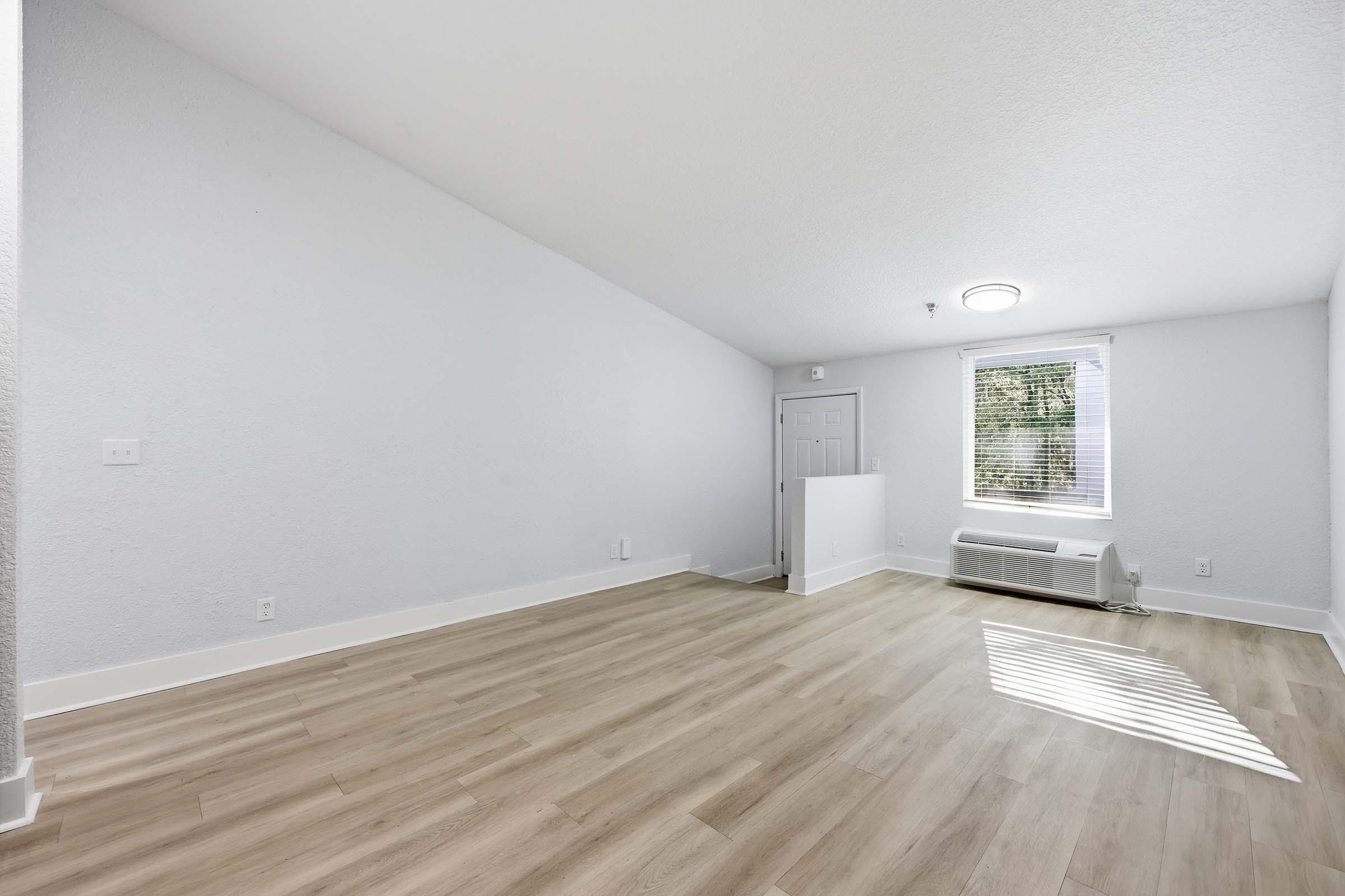 Interior of a light and airy room with high ceilings, featuring beige laminate flooring, a small window with blinds, and a door. The walls are painted white, and an air conditioning unit is visible along one wall. The space is empty, creating a clean, minimalist aesthetic.
