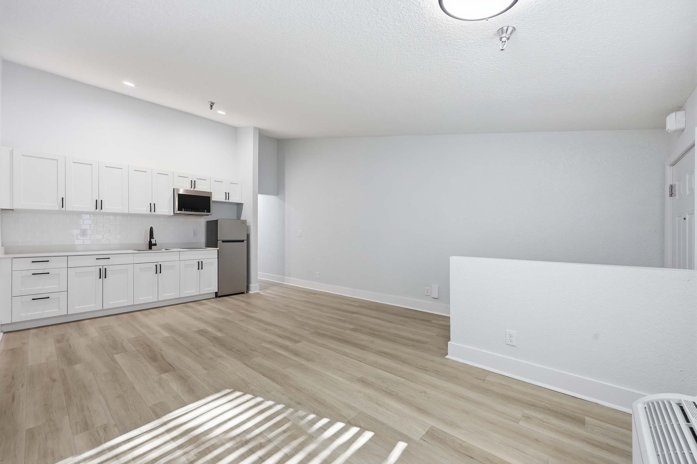 A bright, modern studio apartment with a light wood floor. The left side features a white kitchenette with cabinets, a microwave, and a stainless steel refrigerator. The right side has a spacious area with minimal furnishings and light streaming in through a nearby window. The walls are painted a soft gray.