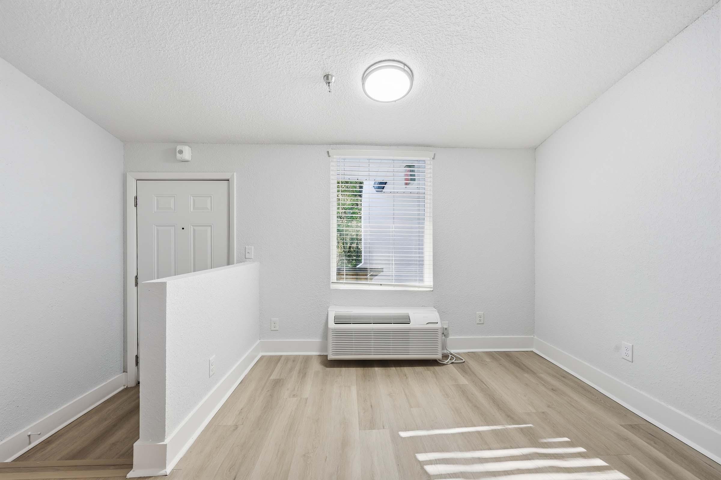 Interior view of a bright, minimally furnished room featuring light-colored walls and wooden flooring. A window allows natural light to enter, and there's an air conditioning unit installed. The space is clean and empty, suitable for various uses.