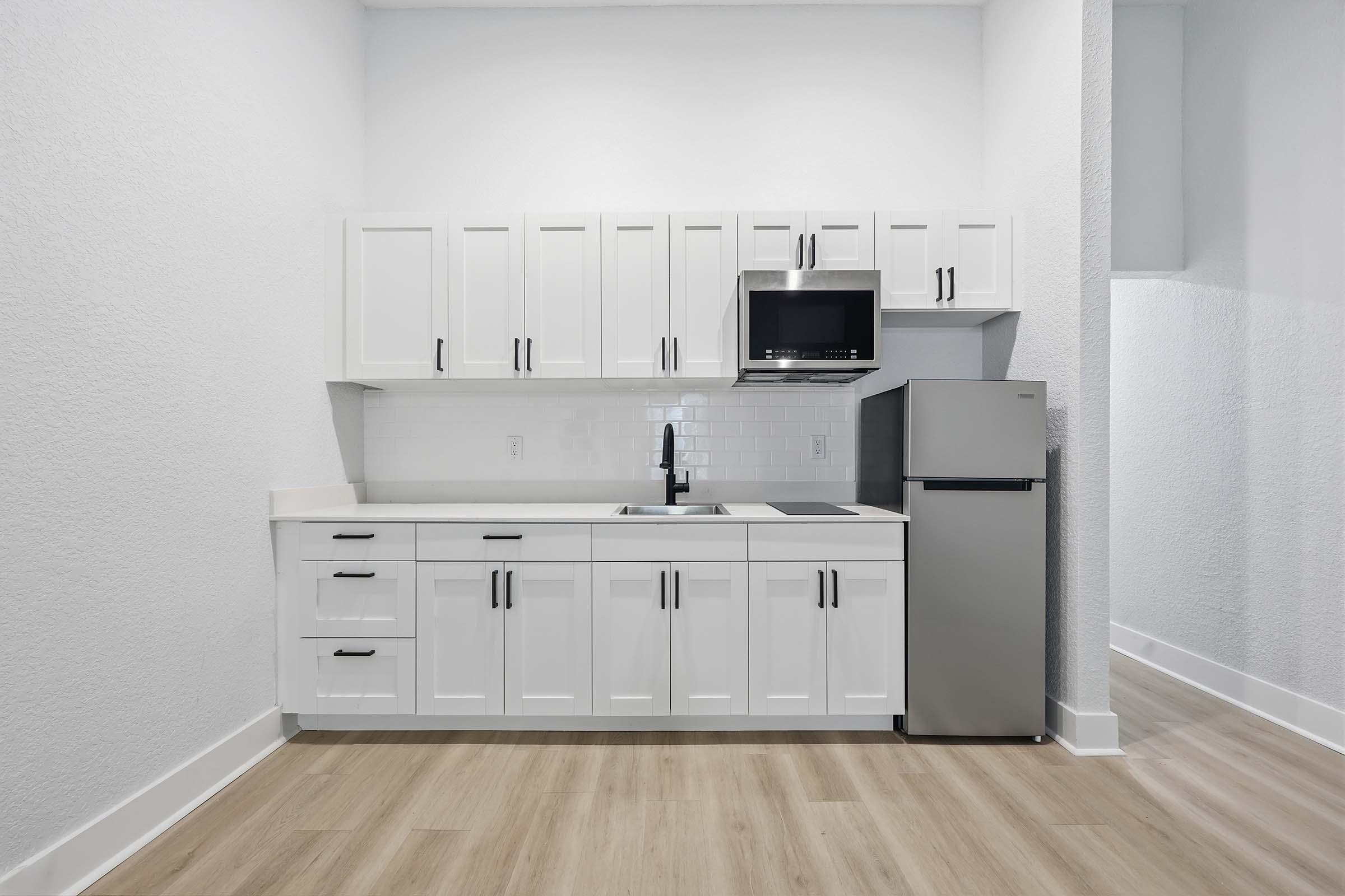 Modern kitchen setup featuring white cabinetry with sleek black handles, a stainless steel refrigerator, a compact black microwave mounted above the countertop, and a minimalist sink area. The space has light-colored walls and a wood-like floor, creating a bright and open atmosphere.