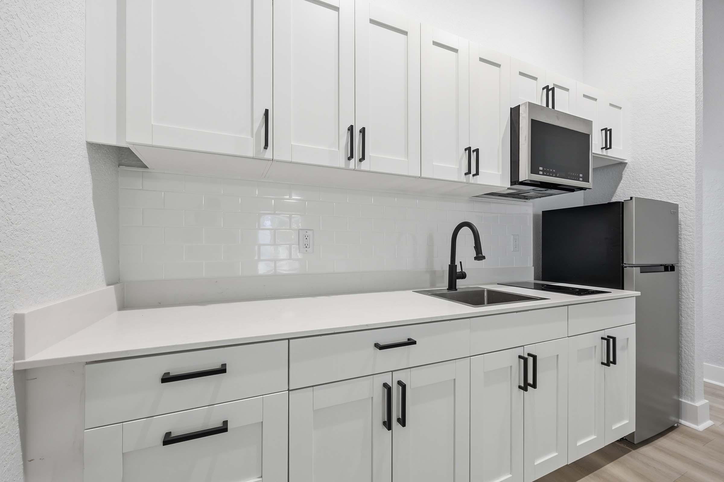 A modern kitchen featuring white cabinets with black handles, a stainless steel microwave, and a sleek sink. The countertop is light-colored, and there's a black refrigerator. The backsplash consists of glossy white tiles, creating a clean and contemporary look.