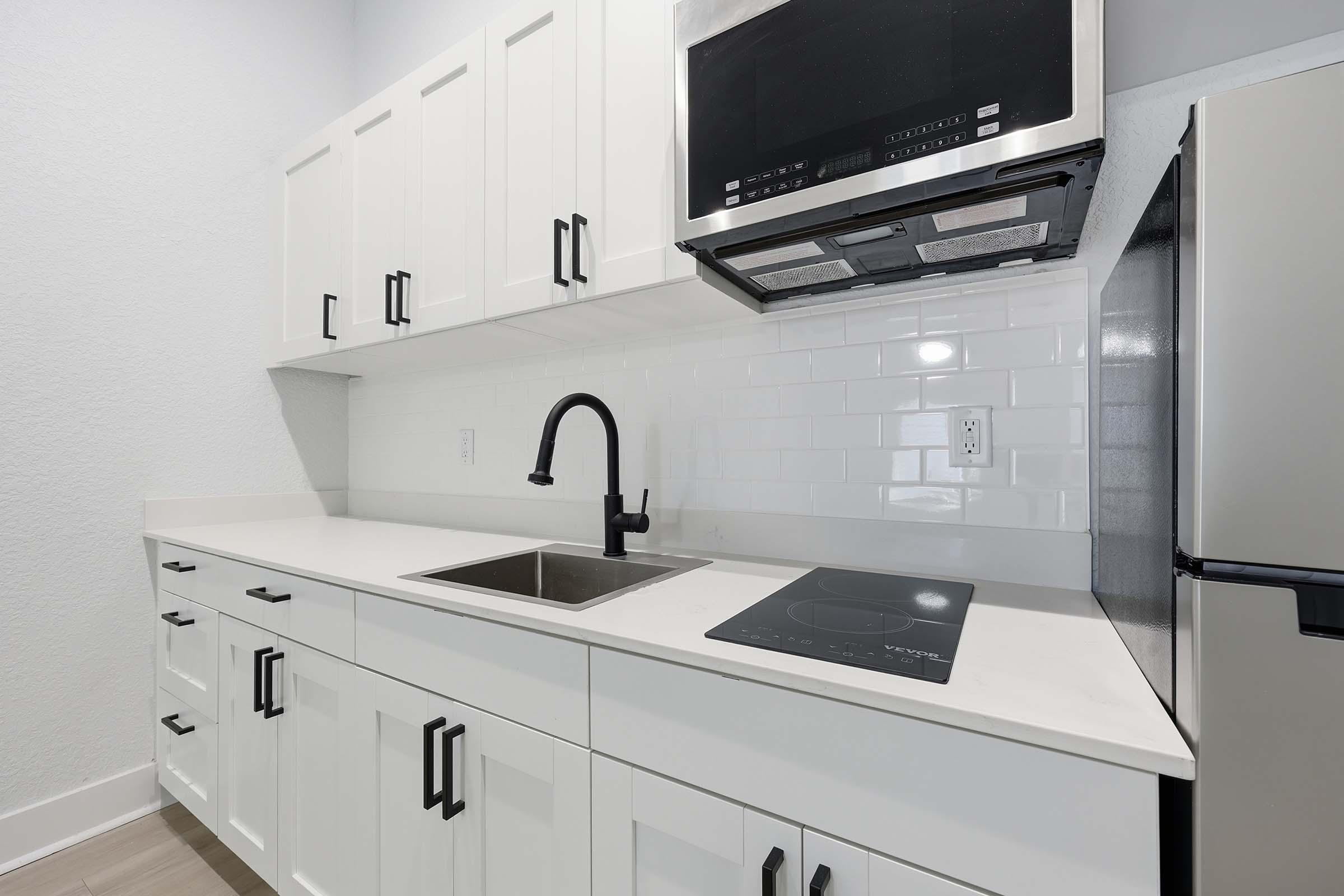 A modern kitchen featuring white cabinetry with black handles, a stainless steel refrigerator, a built-in microwave above a cooktop, and a sleek sink. The countertops are light-colored with a minimalist design, complemented by white subway tile backsplash and neutral walls.