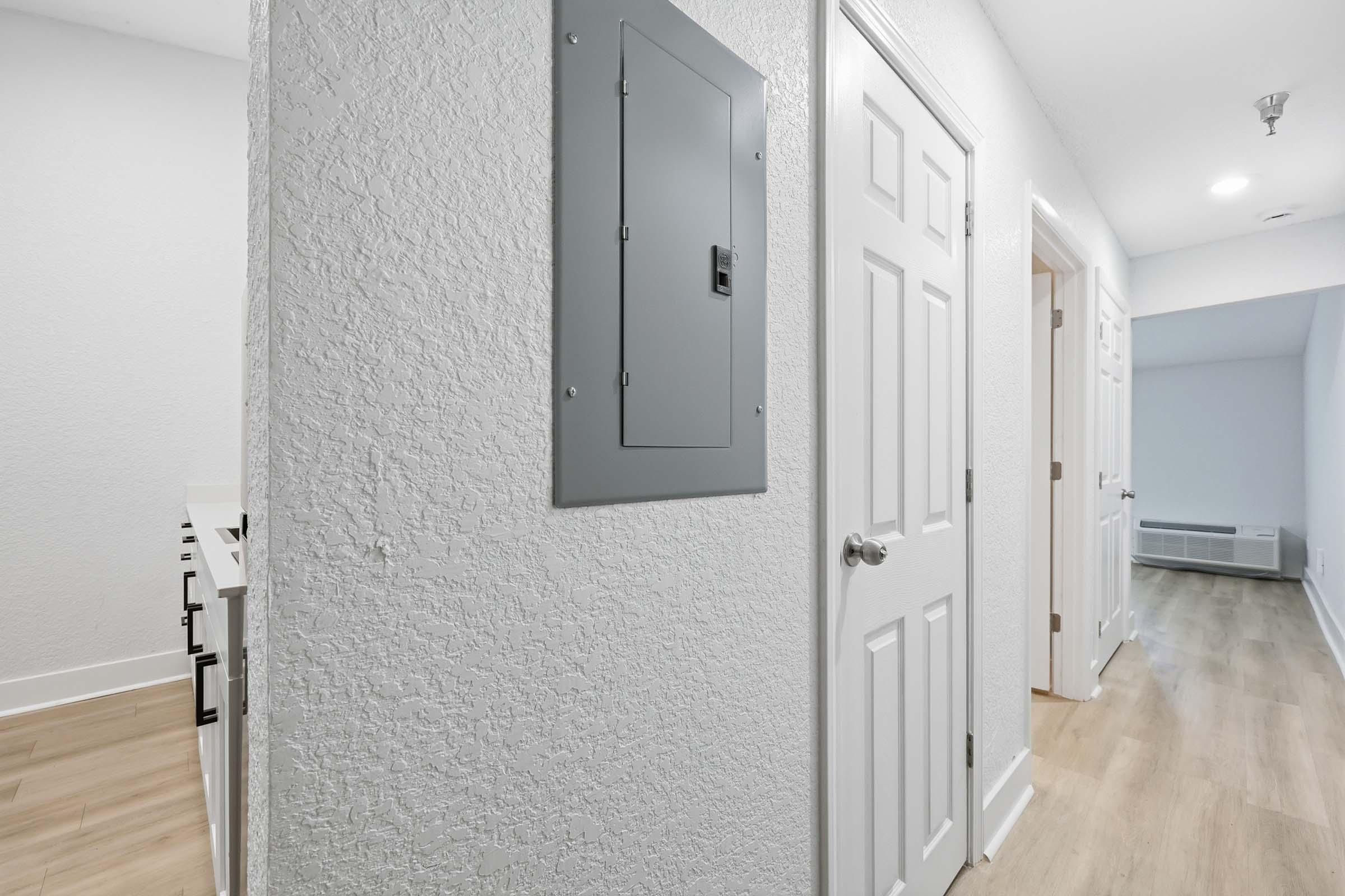 A hallway in a modern interior with light-colored walls and wooden flooring. An electrical panel is mounted on the wall to the left, and a closed door is visible nearby. At the end of the hallway, another area is slightly visible, creating an open feel to the space.