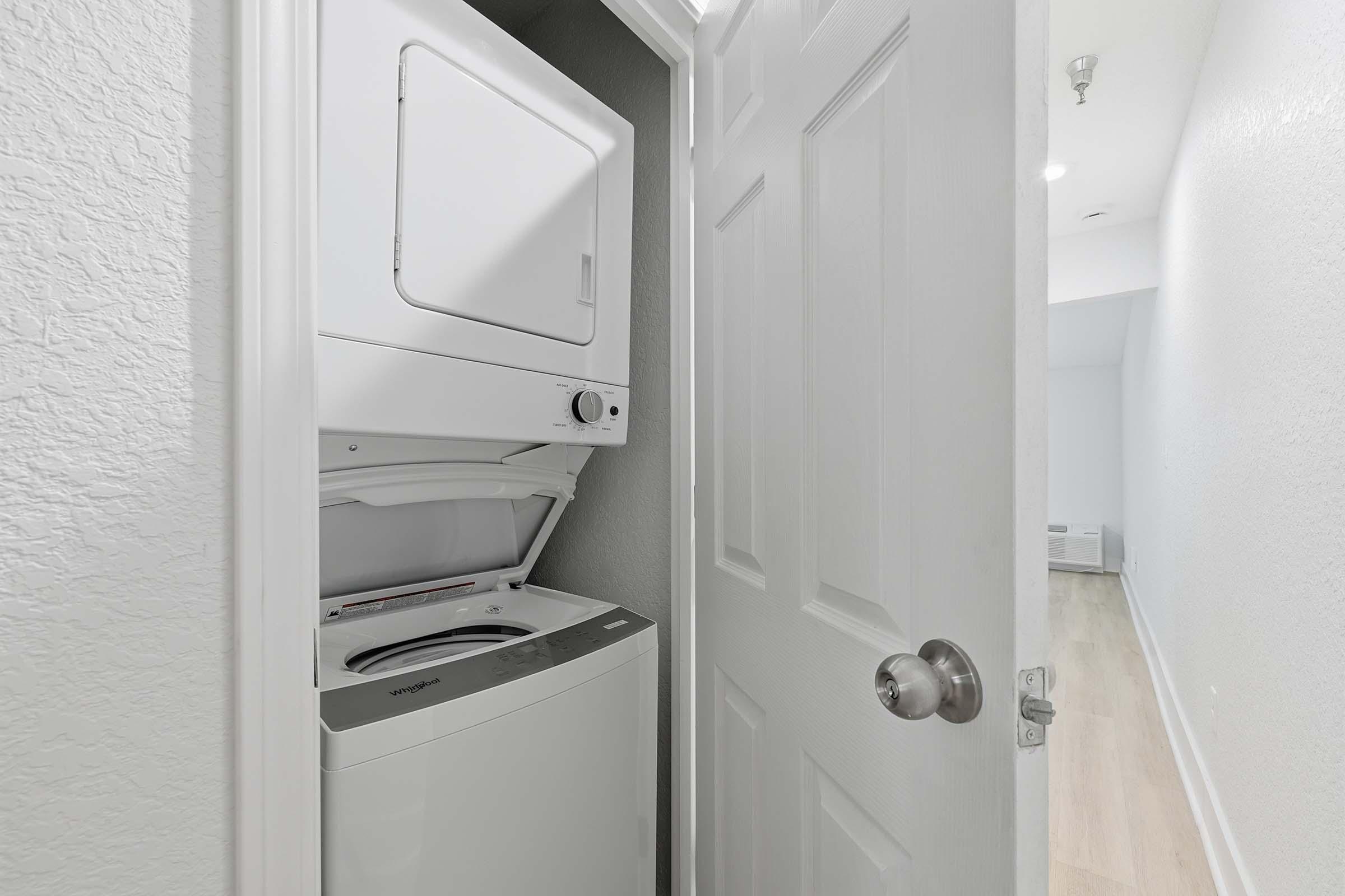 A compact laundry area featuring a stacked washer and dryer unit, positioned against a light-colored wall. The door is open, revealing the appliances, with wooden flooring visible in the background, leading into a well-lit, empty room.