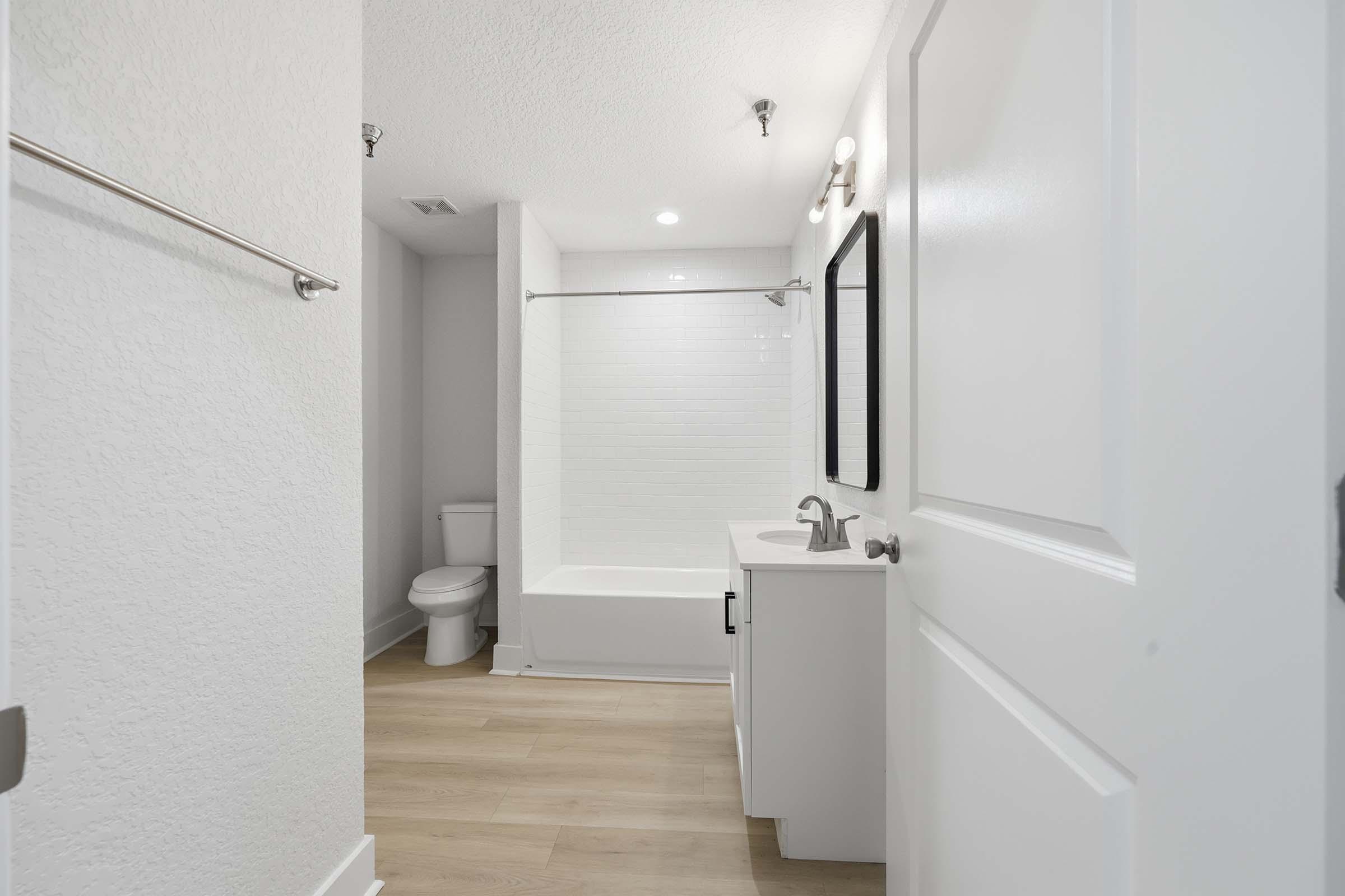 A modern bathroom featuring a bathtub with a shower, a white vanity with a sink, a large mirror, and a toilet. The walls are light-colored, and the floor has a wooden appearance, creating a clean and bright atmosphere.