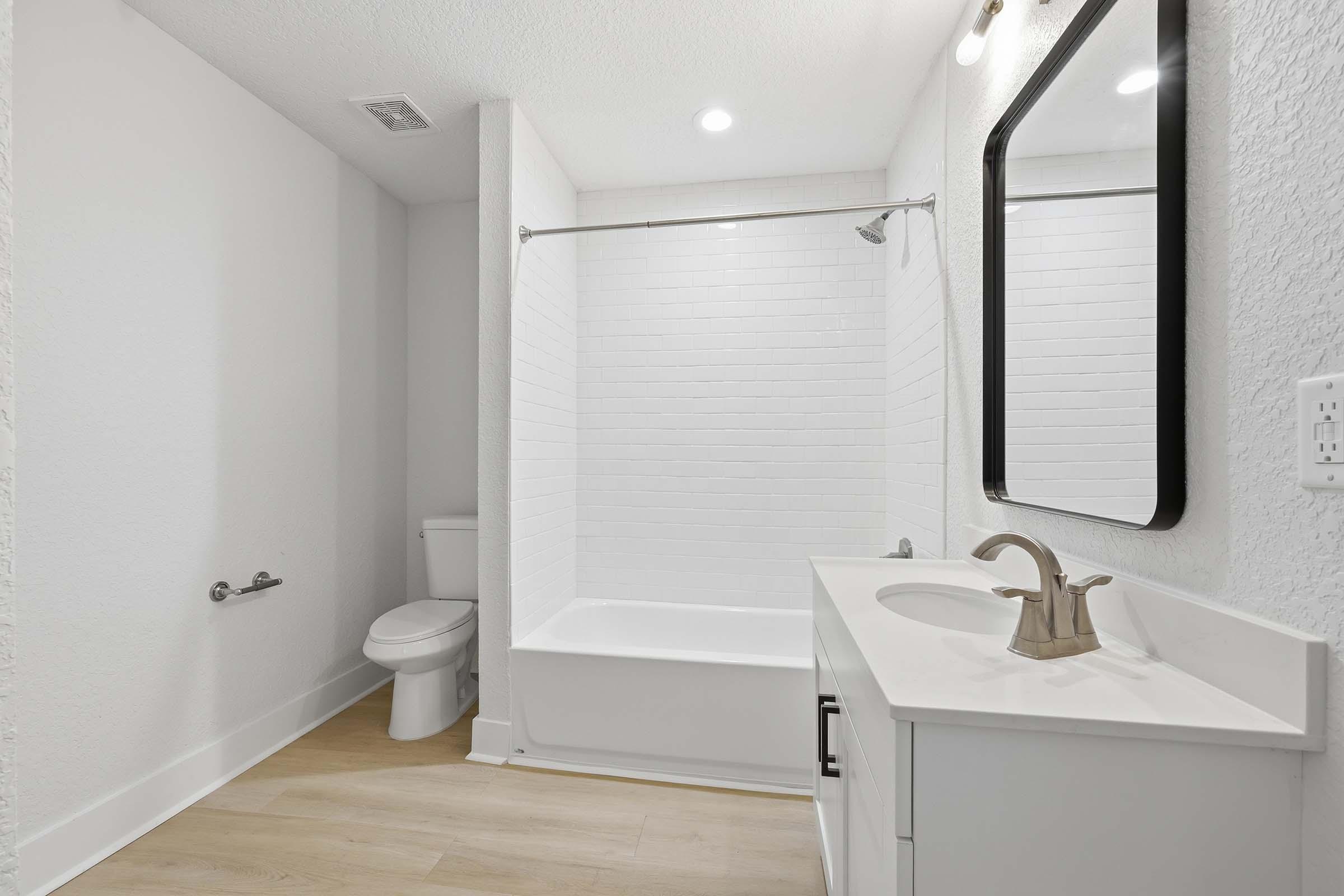 A modern bathroom featuring a white bathtub with a shower, a wall-mounted mirror above a white vanity with a sink, and neutral-colored walls. The space is well-lit with overhead lights and has light wood flooring. A toilet is positioned next to the tub, completing the clean and minimalist design.