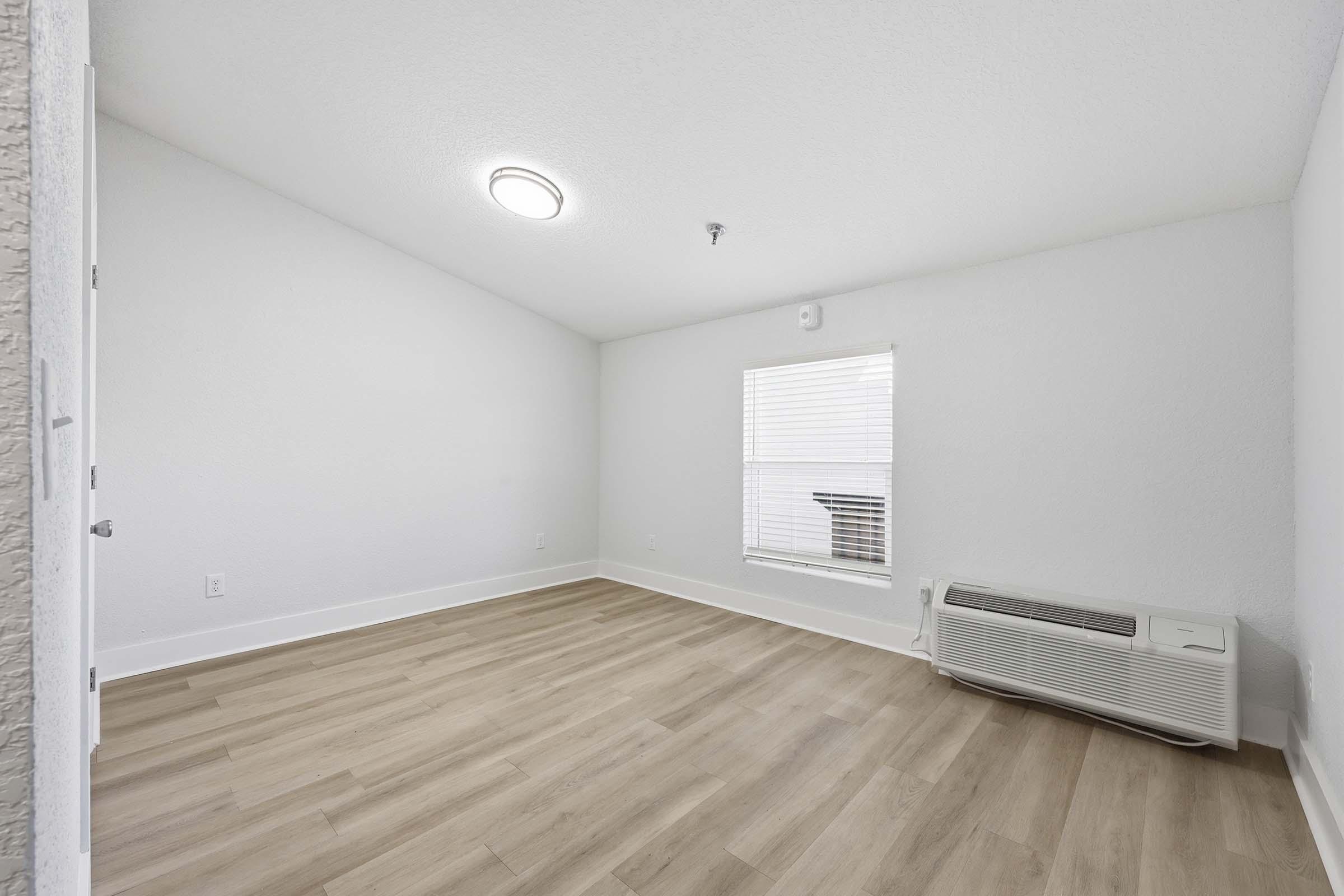 A bright, empty room with light-colored walls and hardwood flooring. There is a window with white blinds allowing natural light to enter, and an air conditioning unit along one wall. The ceiling is slightly sloped with a modern ceiling light fixture. The overall atmosphere is clean and minimalistic.