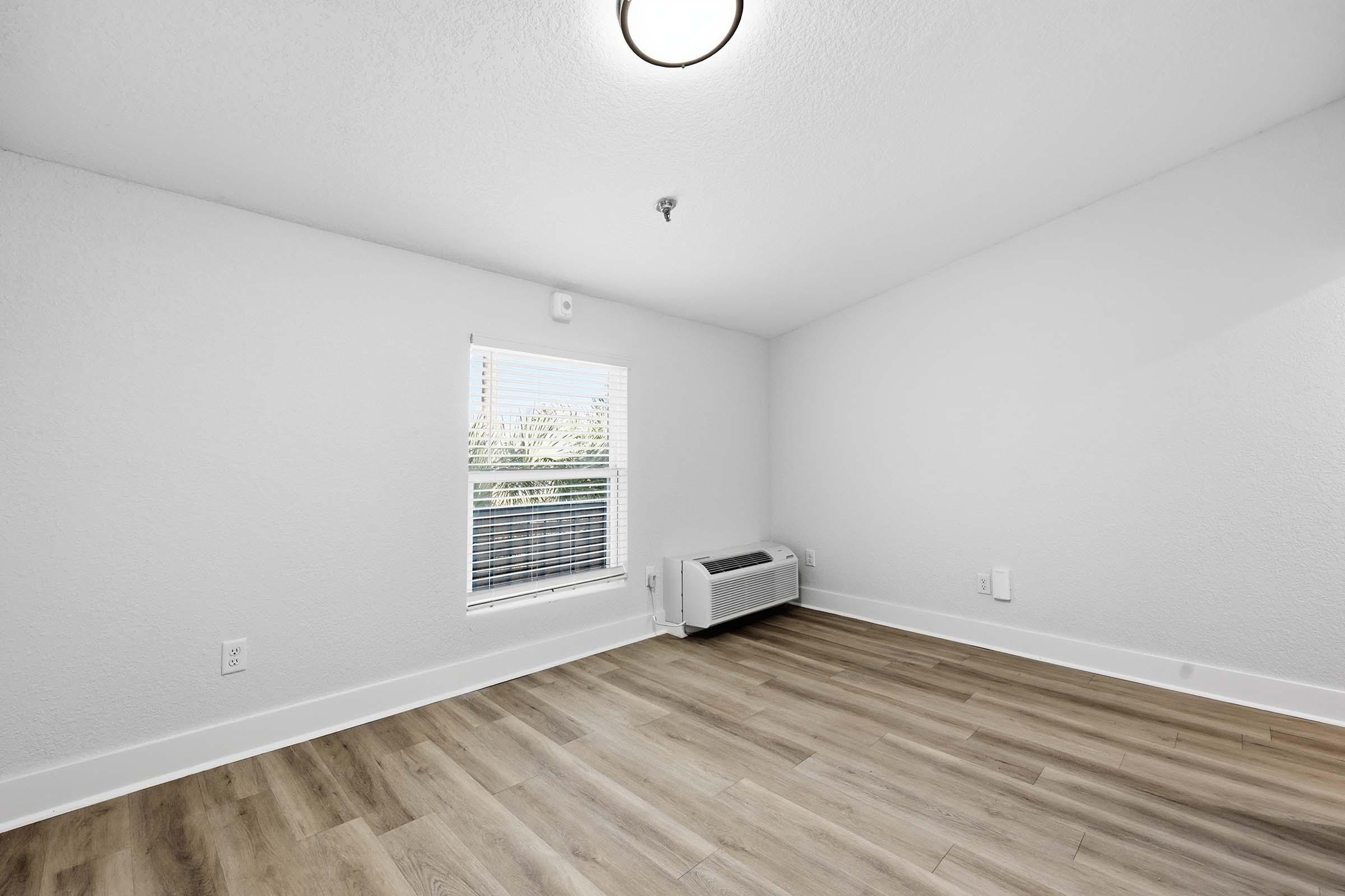 Empty room with light-colored walls and wood-like flooring. A single window lets in natural light, and there is a wall-mounted air conditioning unit below the window. The space is bright and minimalist, suitable for various uses.