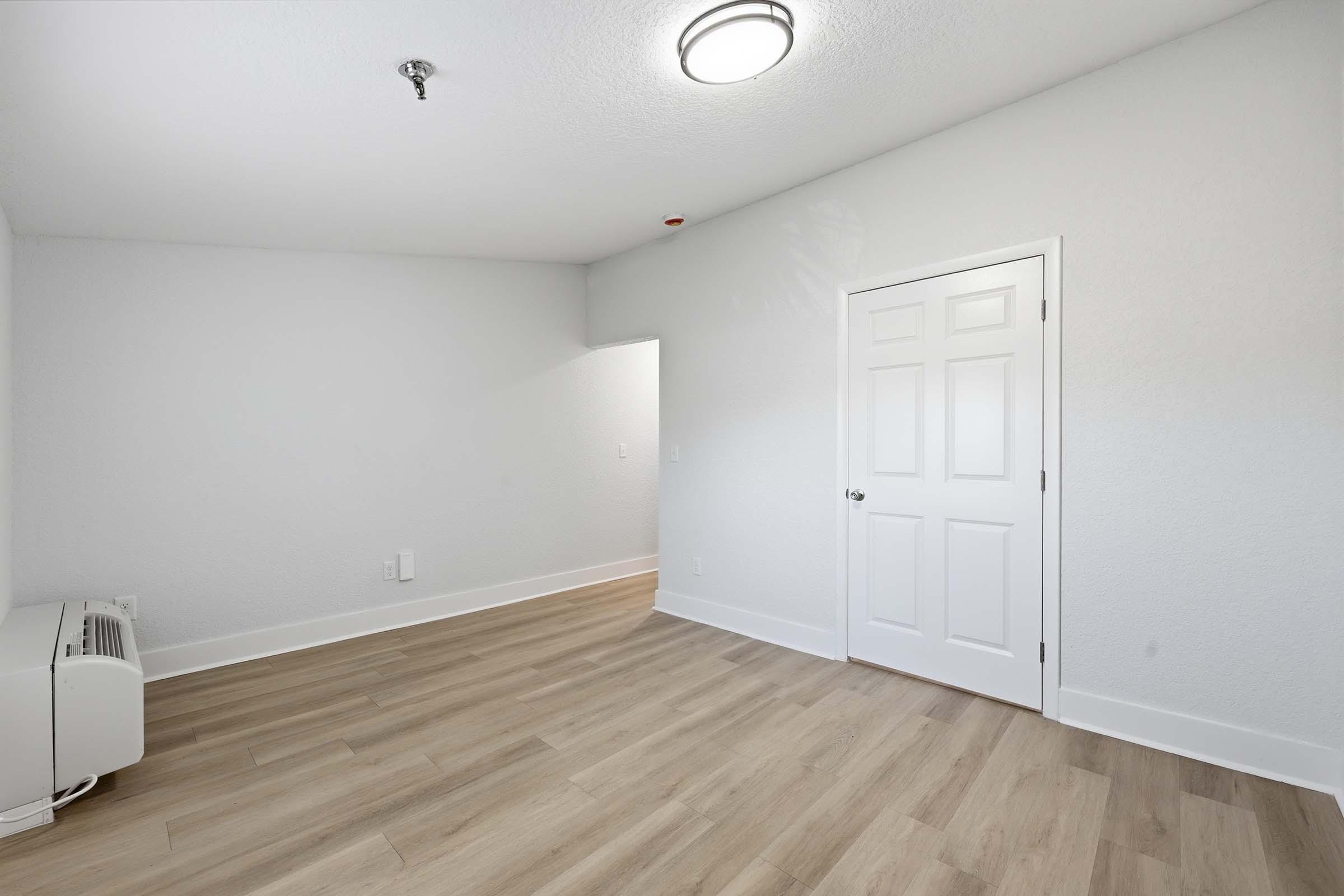 A well-lit, empty room with light-colored walls and laminate flooring. There is a small air conditioning unit on the left and a closed white door on the right. A circular ceiling light illuminates the space, which has a modern, clean look.