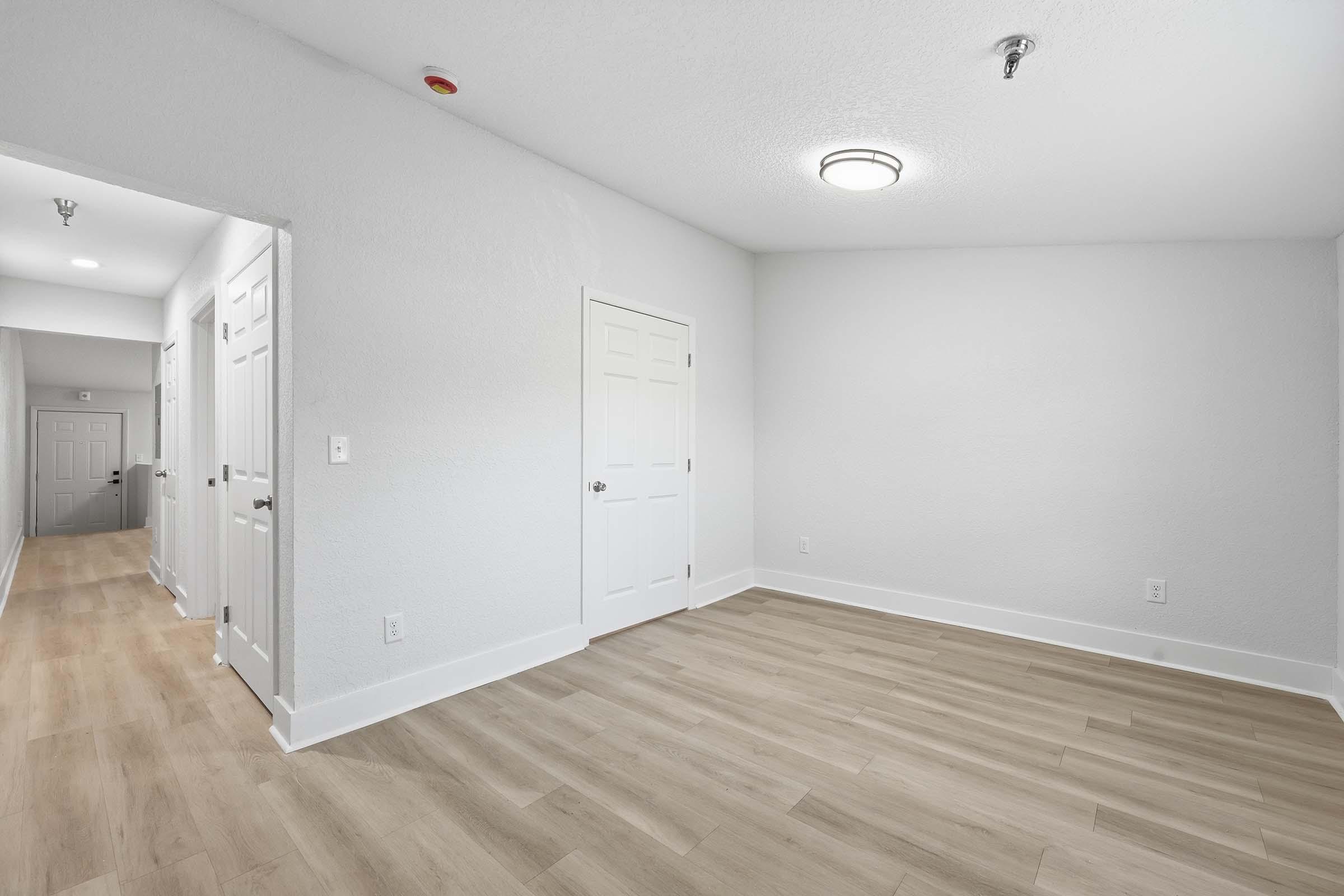 A bright, empty room with light gray walls and wood-like flooring. Doors are visible on the left and a ceiling light fixture is installed. The space appears clean and modern, suitable for various uses like a living area or office.