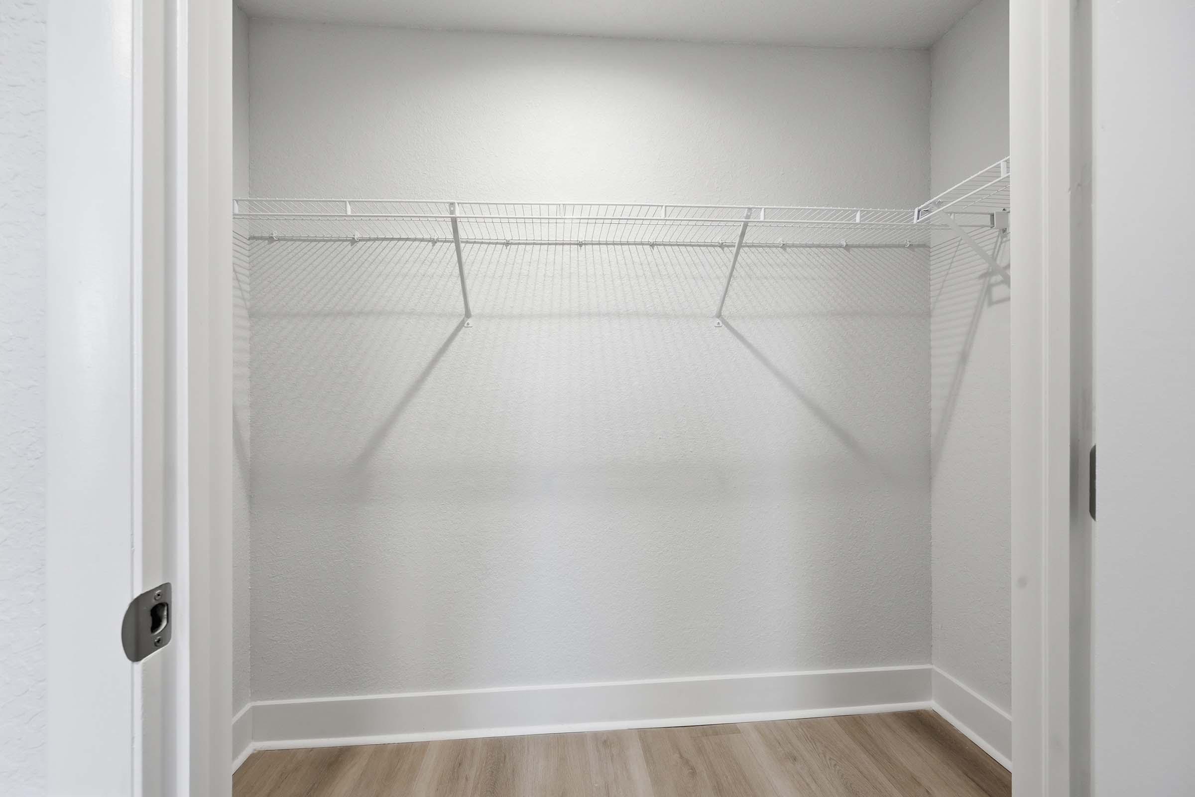 An empty closet with light gray walls, featuring a wire shelf mounted on the back wall. The floor is light wood, and there is a white door partially visible on the left side of the frame.