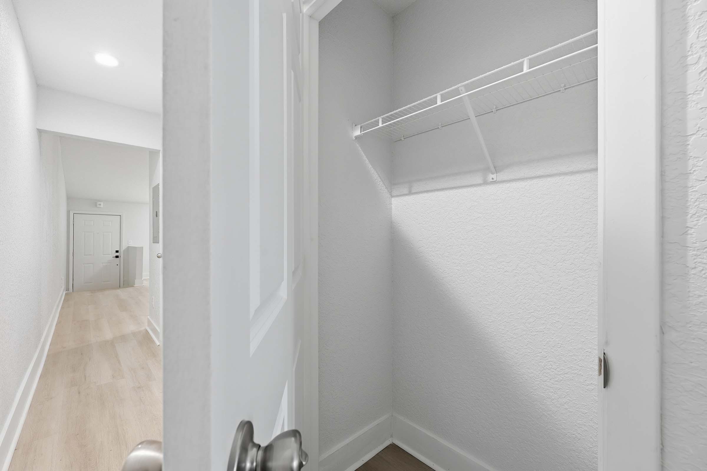 A view into a small, empty closet with a shelf, surrounded by light-colored walls and a wooden floor. The door is partially open, revealing a well-lit hallway beyond with another door visible in the distance.
