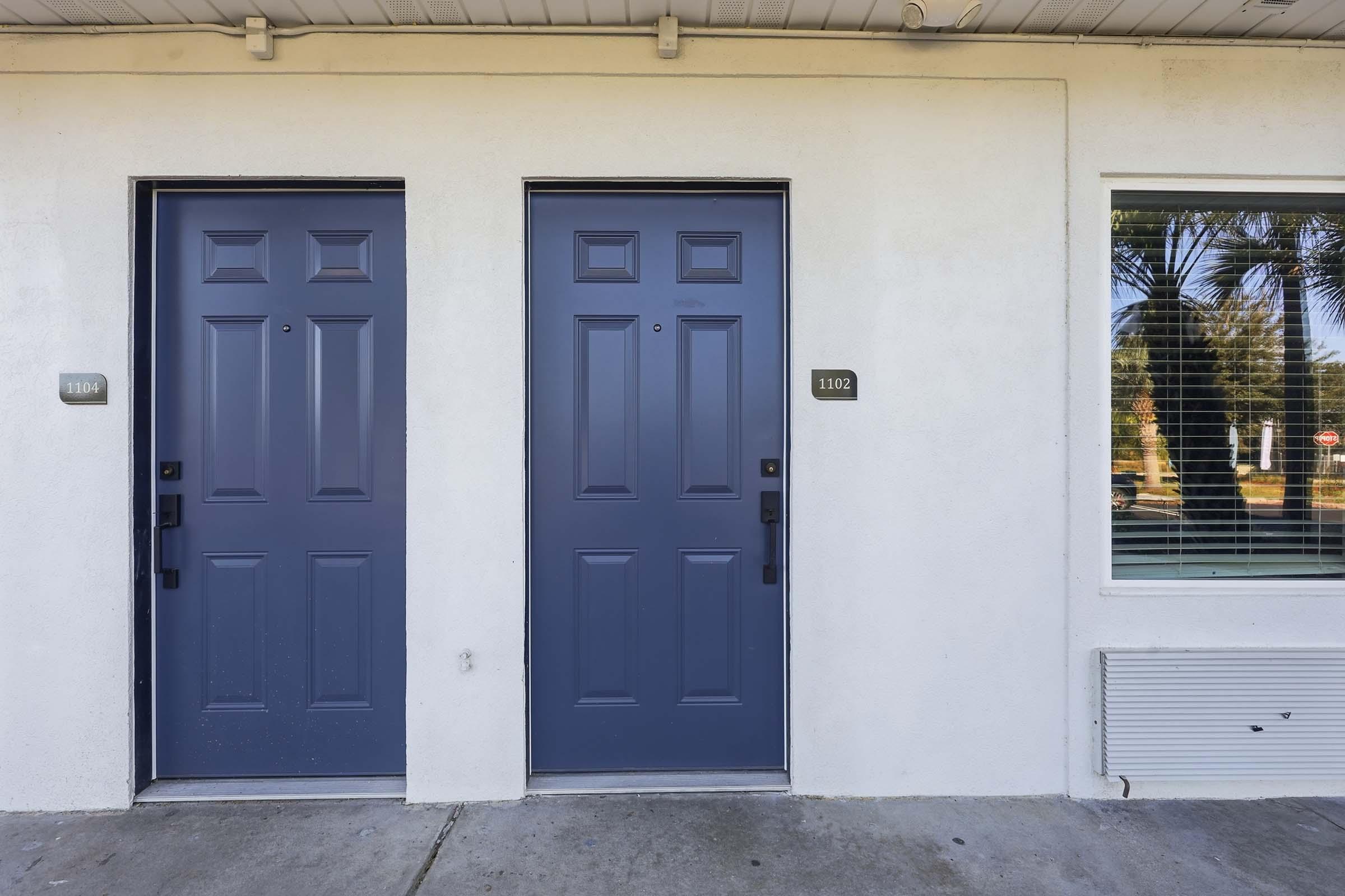 Two blue doors side by side on a white wall, with door numbers 1102 and 1103. The doors have black handles and a small window adjacent to them, showcasing blinds and a view of palm trees outside. The setting appears to be a residential or commercial building entrance.