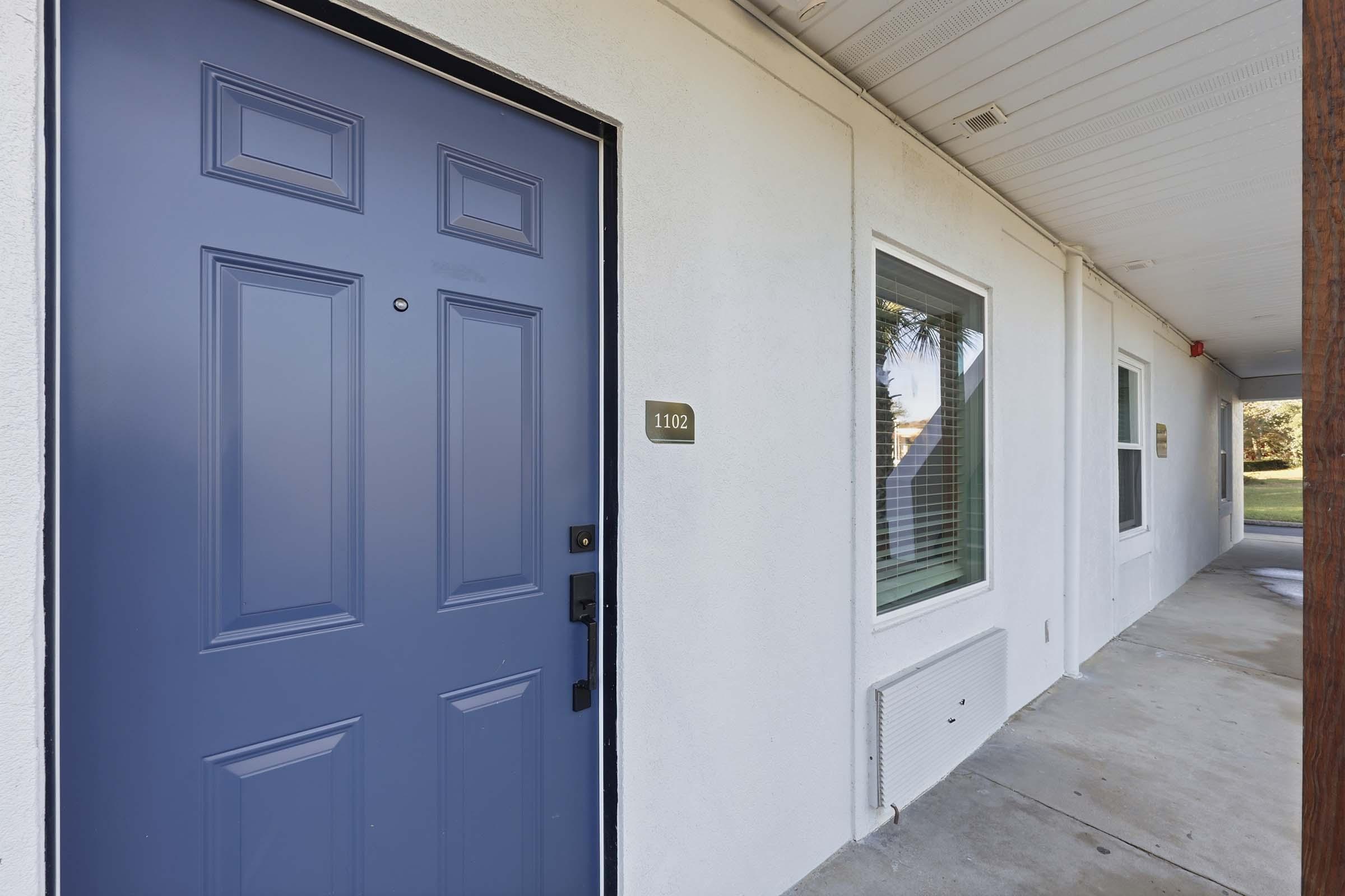 A blue door labeled "1102" is the focus of the image, set against a white wall. There are windows next to the door, providing a view of the exterior. The ground is concrete, and the entrance has a covered area, creating a welcoming appearance.