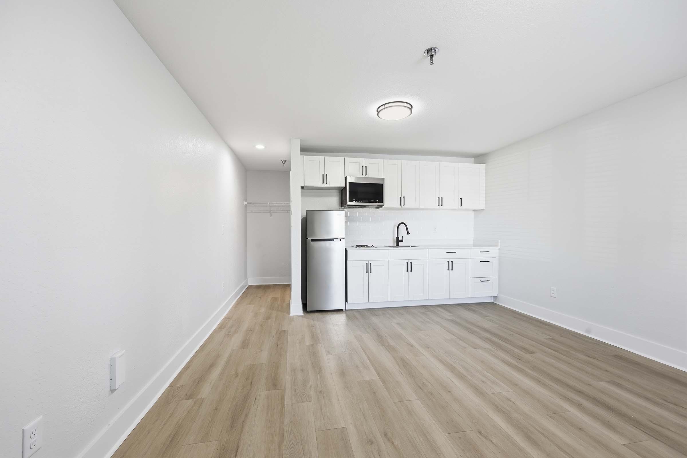 Modern kitchen and living area in a bright, minimalist apartment. The space features light wood flooring, white cabinetry, stainless steel refrigerator, and microwave. Natural light fills the room through windows, creating an open and airy atmosphere.