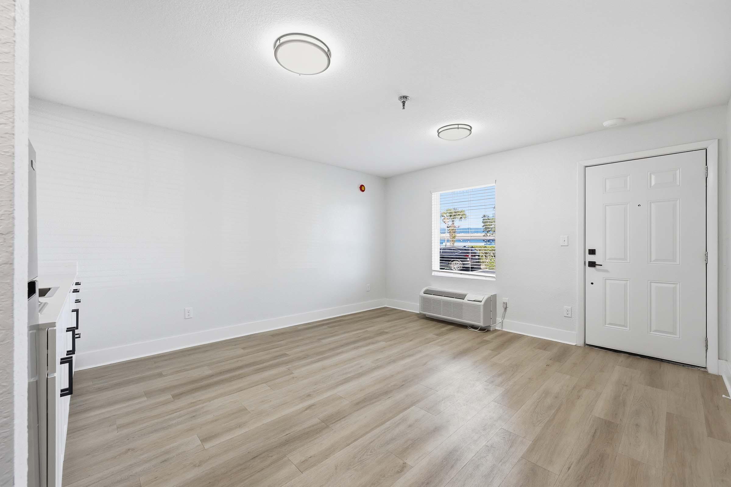 A spacious, well-lit room with light-colored walls and wooden flooring. Two ceiling lights provide illumination. A window on the left lets in natural light, and there's an air conditioning unit beneath it. The door on the right leads outside, completing the simple and modern aesthetic of the space.
