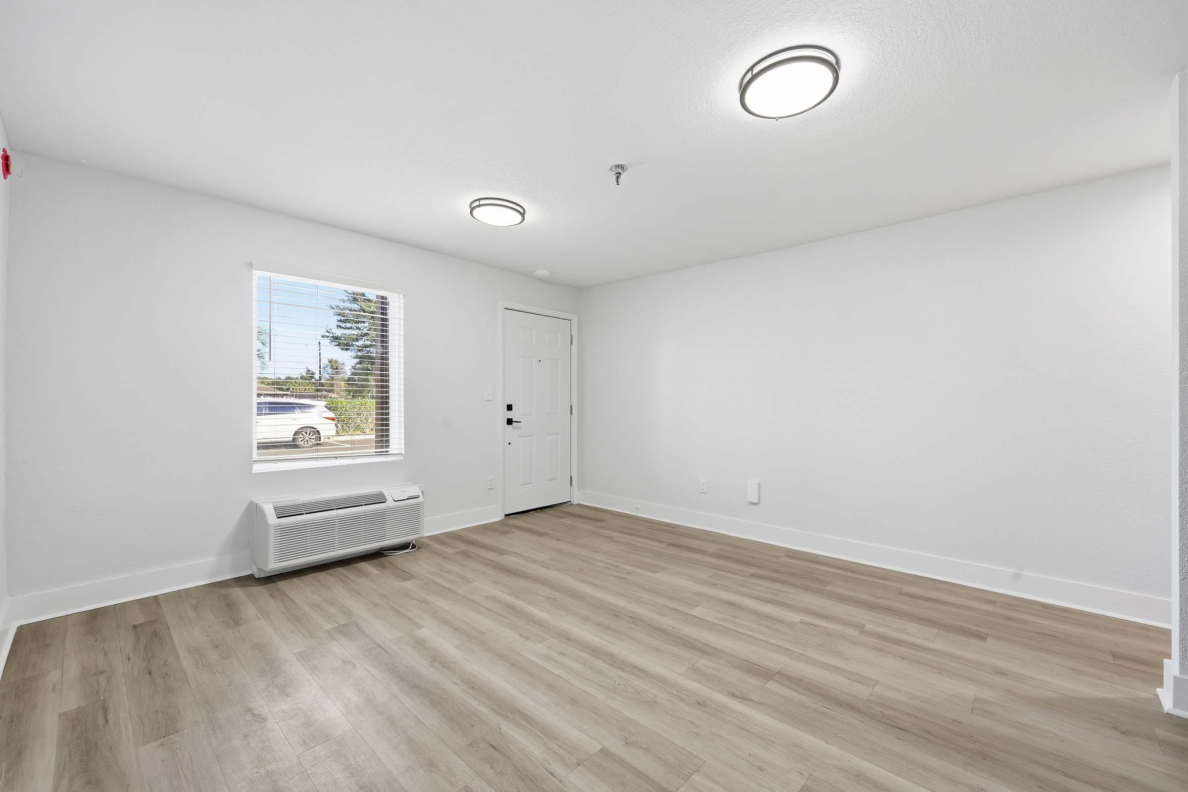 A bright, empty interior room featuring light-colored walls and modern light fixtures. The flooring is a light wood style, and there is a large window allowing natural light. An air conditioning unit is visible on the wall below the window. The entrance door is centered on the wall, adding to the open feel of the space.