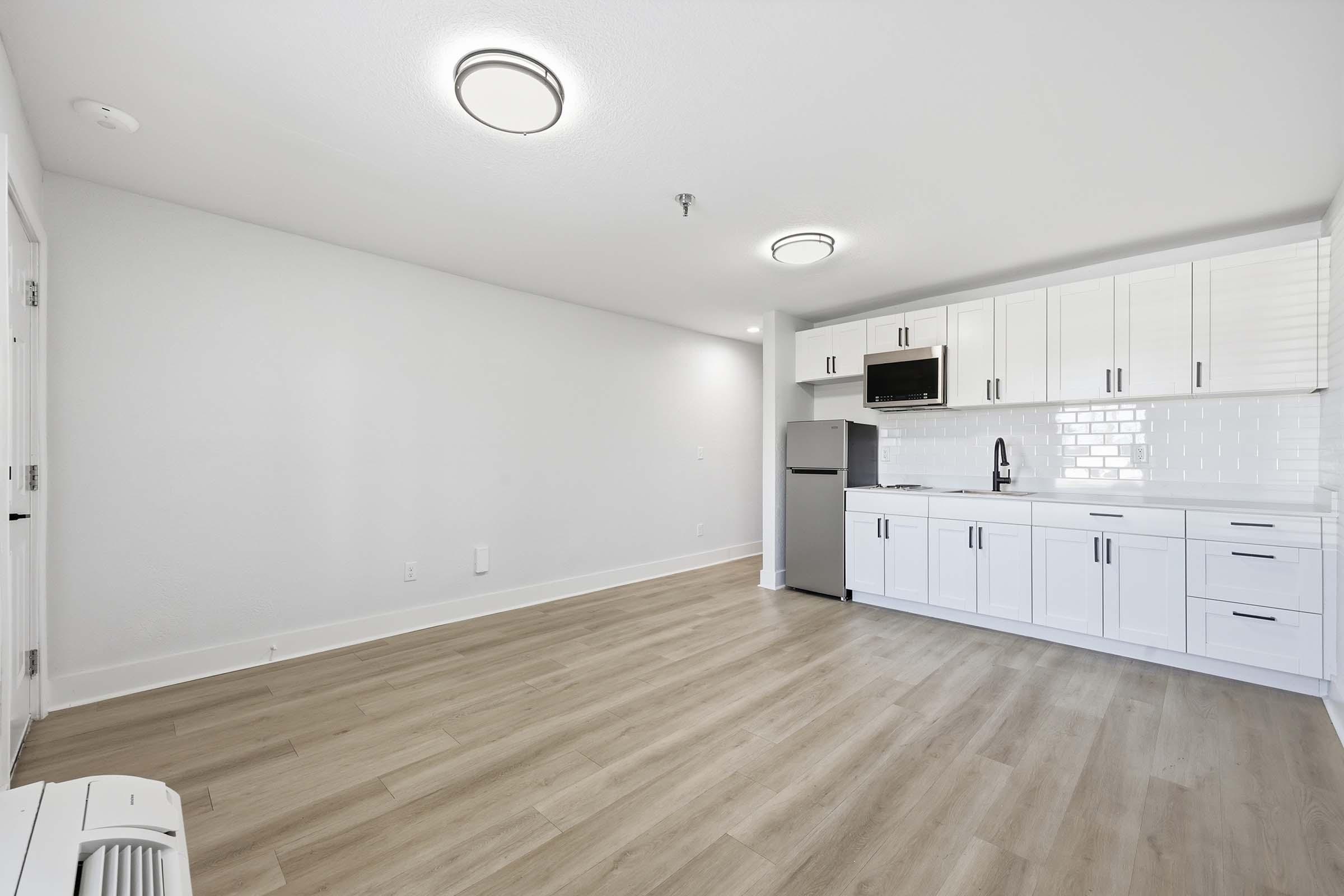 Modern, minimalist kitchen and living space featuring light wood flooring, white cabinetry, and stainless steel appliances. The kitchen area includes a refrigerator, microwave, and sink, with white tiles as a backsplash. Bright overhead lighting enhances the spacious, clean atmosphere.
