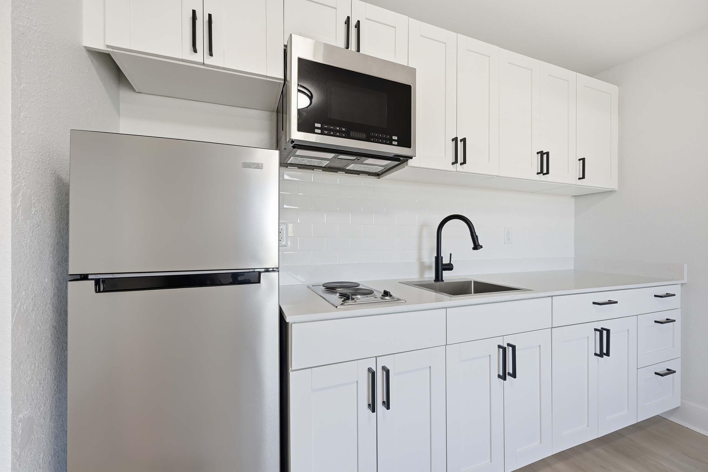 A modern kitchen featuring white cabinets, a stainless steel refrigerator, a microwave mounted above the stove, and a black faucet over a sink. The countertop is simple, and there is a gas stove with a pan on it. The overall design is clean and contemporary, with a bright and spacious feel.
