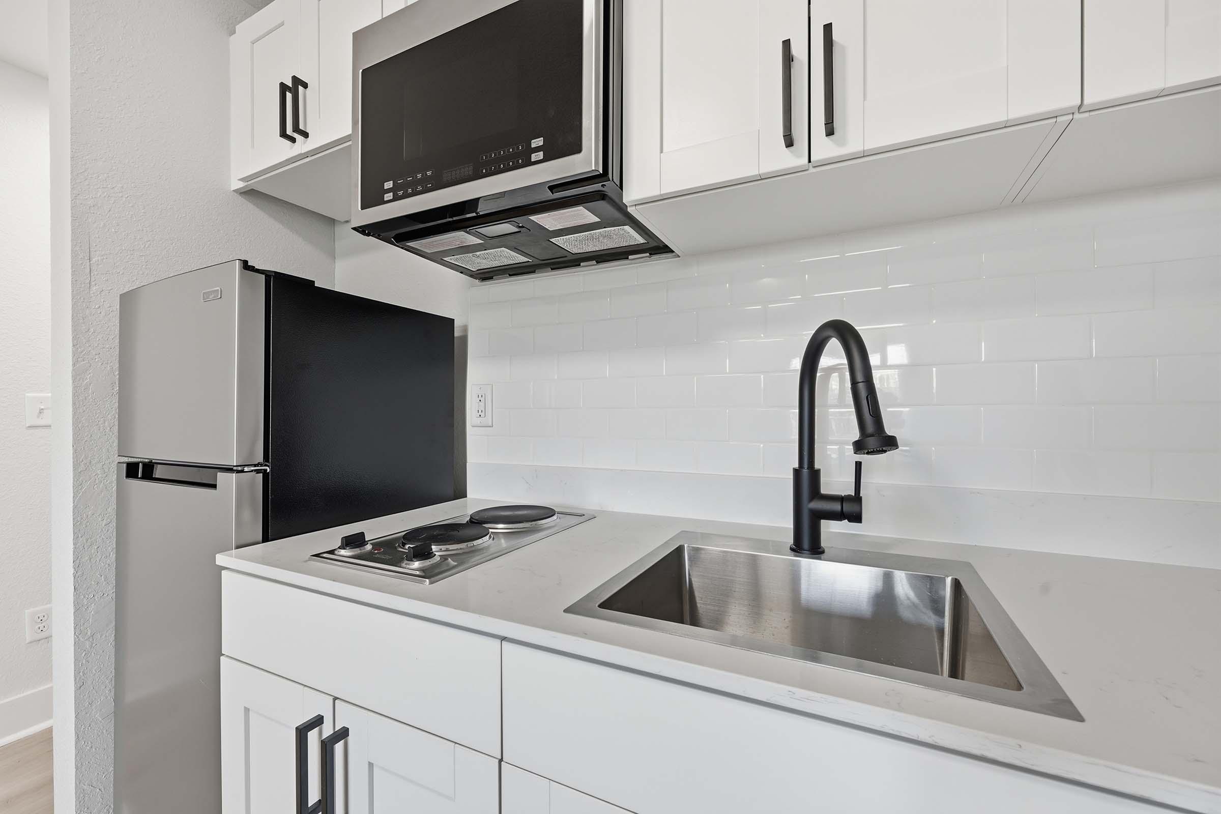 Modern kitchen featuring white cabinets, a stainless steel sink with a black faucet, a black microwave mounted above the cooking area, and a sleek black refrigerator. The countertop is light-colored, complementing the white backsplash tiles, creating a stylish and contemporary look.