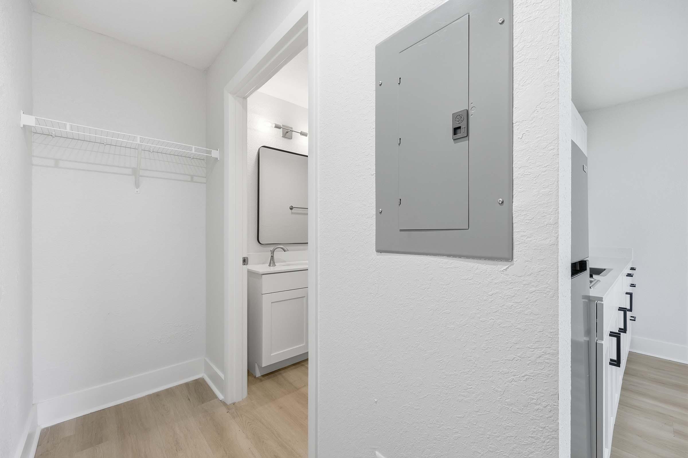 A clean, modern interior view of a small room featuring a white wall with a light switch panel, a utility closet with wire shelving, and a glimpse of a bathroom with a sink and mirror. The flooring is light wood, creating a bright and airy atmosphere.