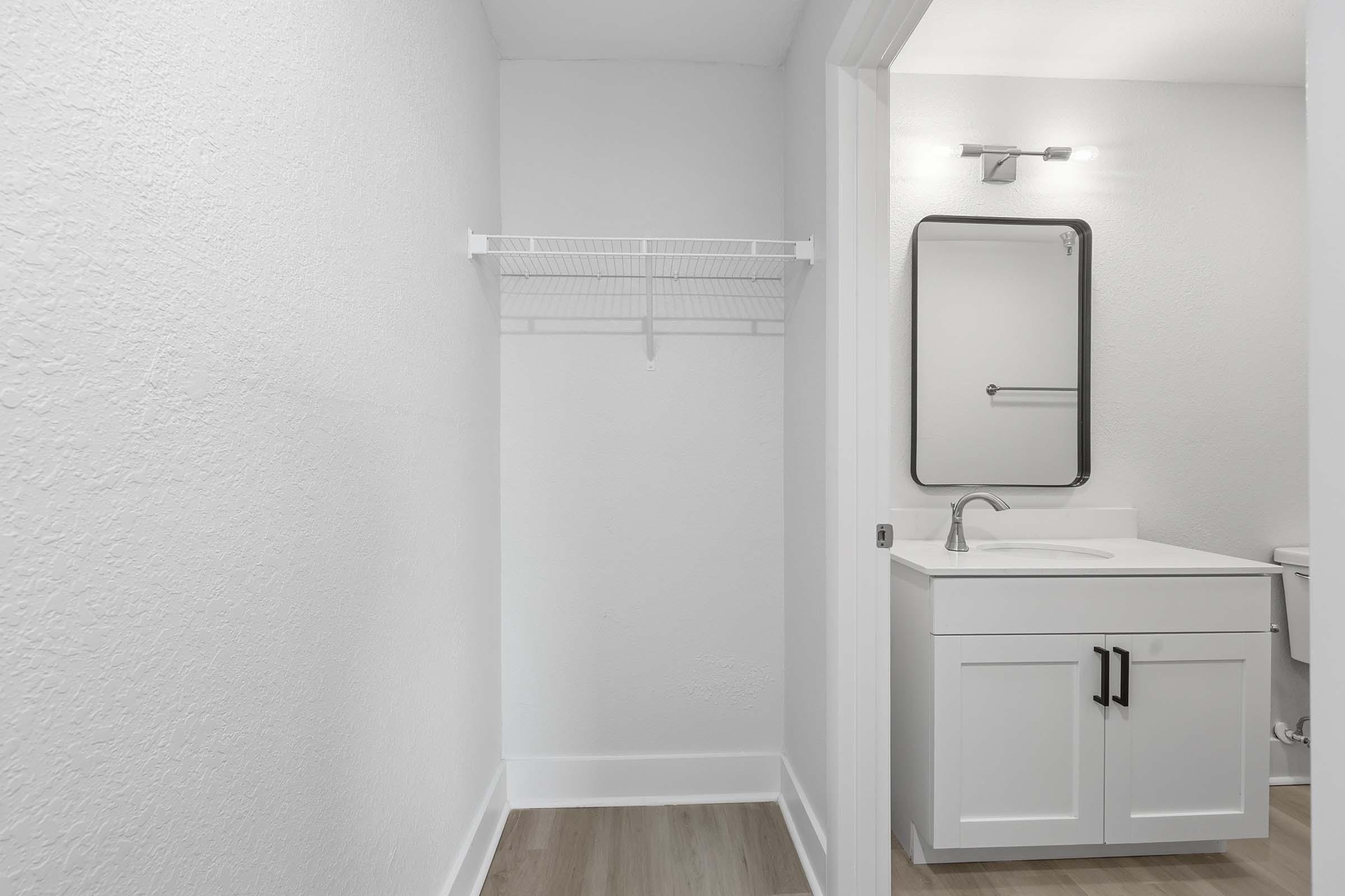 A clean, minimalist bathroom view featuring a white vanity with a sink, a mirror, and a simple light fixture. An empty wall shelf is mounted next to the mirror, and the space has light-colored walls and wooden flooring, creating a bright and modern atmosphere.