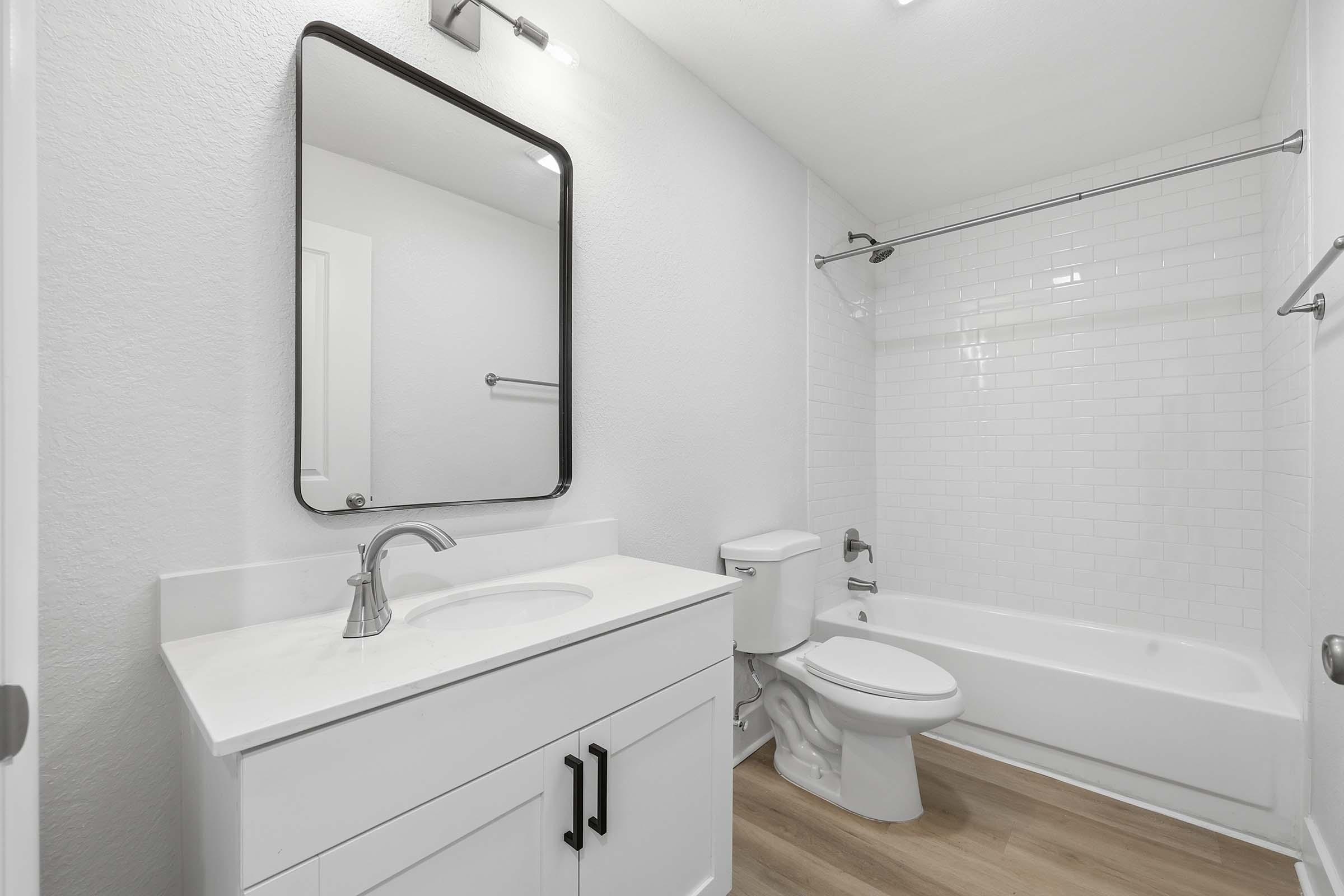 A modern bathroom featuring a white vanity with a sleek sink and faucet, a large mirror above the sink, a bathtub with a shower, and neutral-colored walls. The flooring is a light wood finish, creating a clean and contemporary look. 