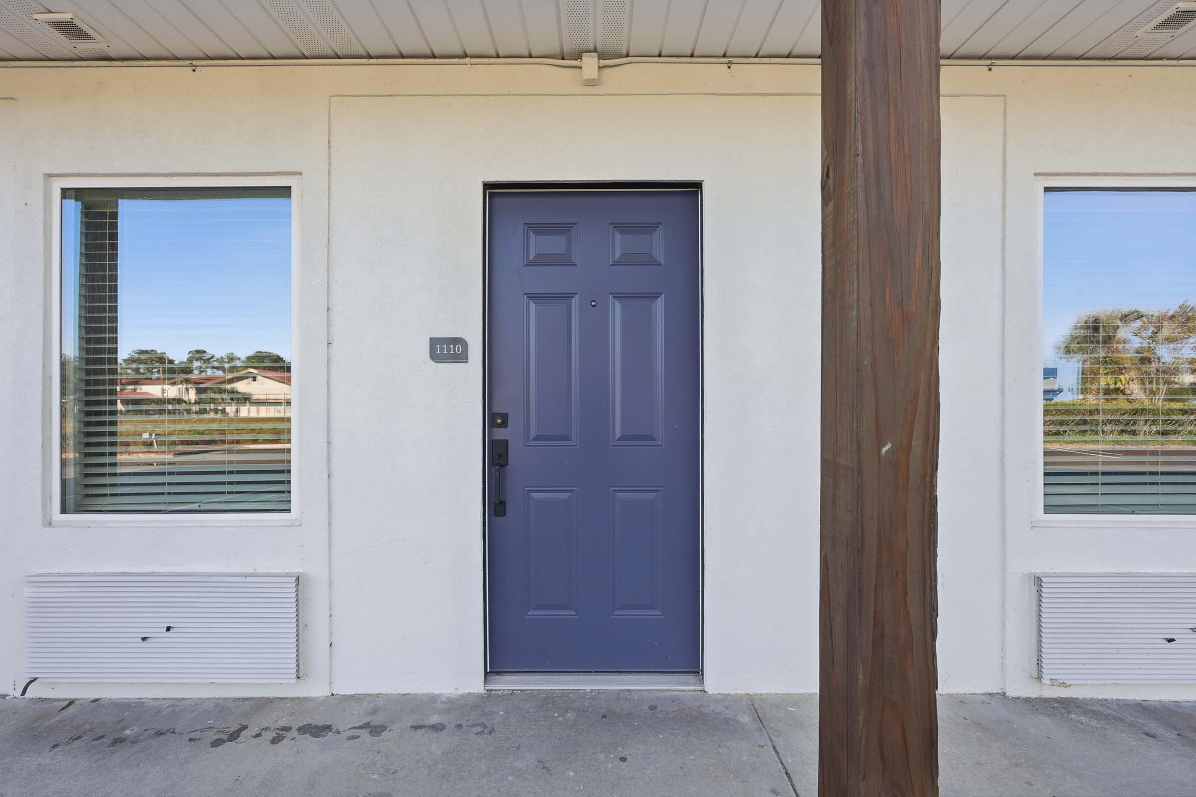 Exterior view of a building featuring a purple front door with the number 1110 on it, flanked by two windows. The walls are white, and there is a wooden post supporting the overhang. Visible outside is a landscaped area, hinting at a sunny day with clear skies.