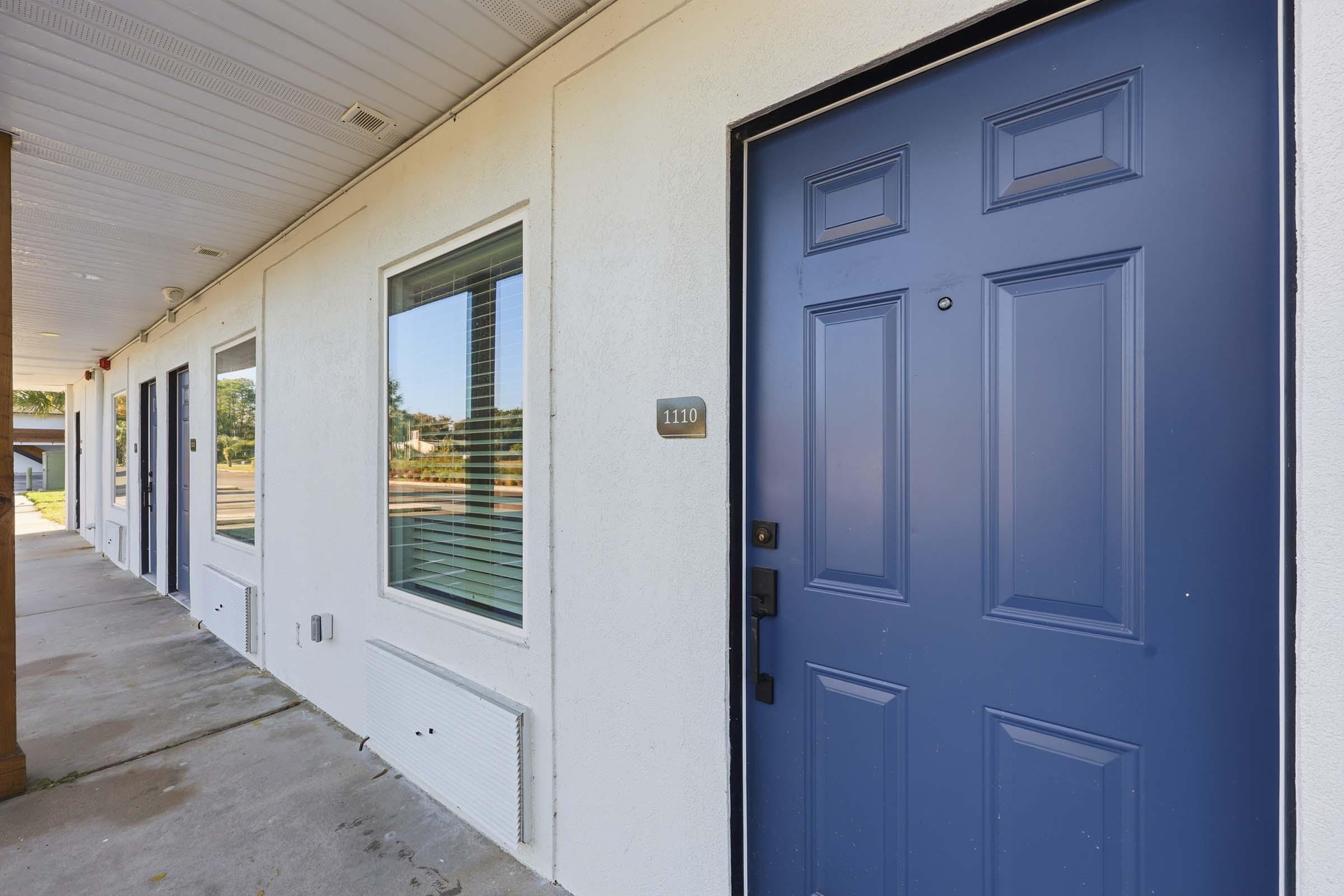 A close-up view of a blue door labeled "1100," located on a building's exterior. The door features a simple design with six raised panels and a black handle. Nearby are two windows with blinds, allowing natural light inside. The setting appears to be part of a well-maintained, single-story building.