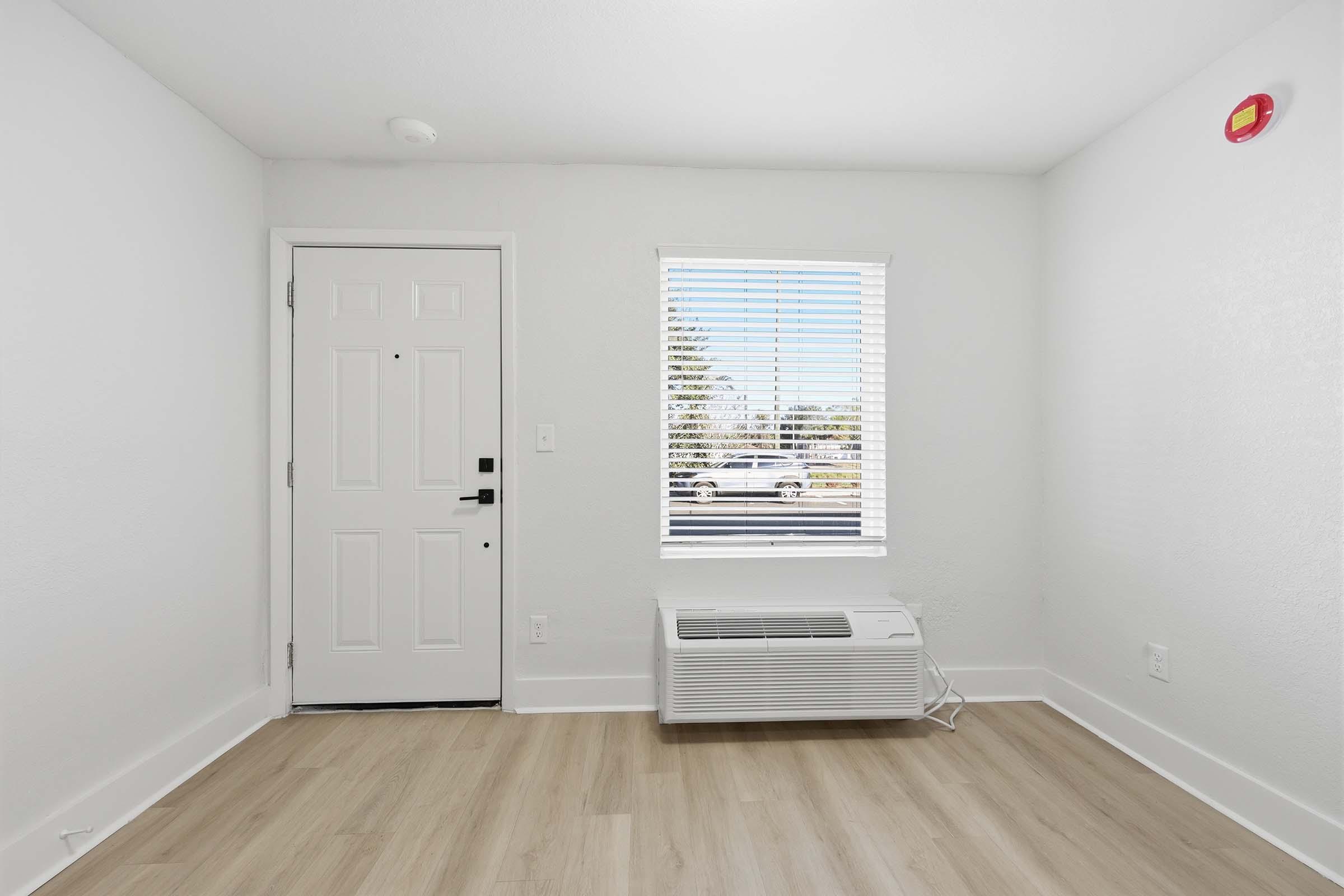 A bright, minimalistic room featuring a white wall, a closed door with a window, and an air conditioning unit on the floor. Natural light streams in through the window with white blinds. The hardwood floor has a light finish, enhancing the spacious feel of the area.
